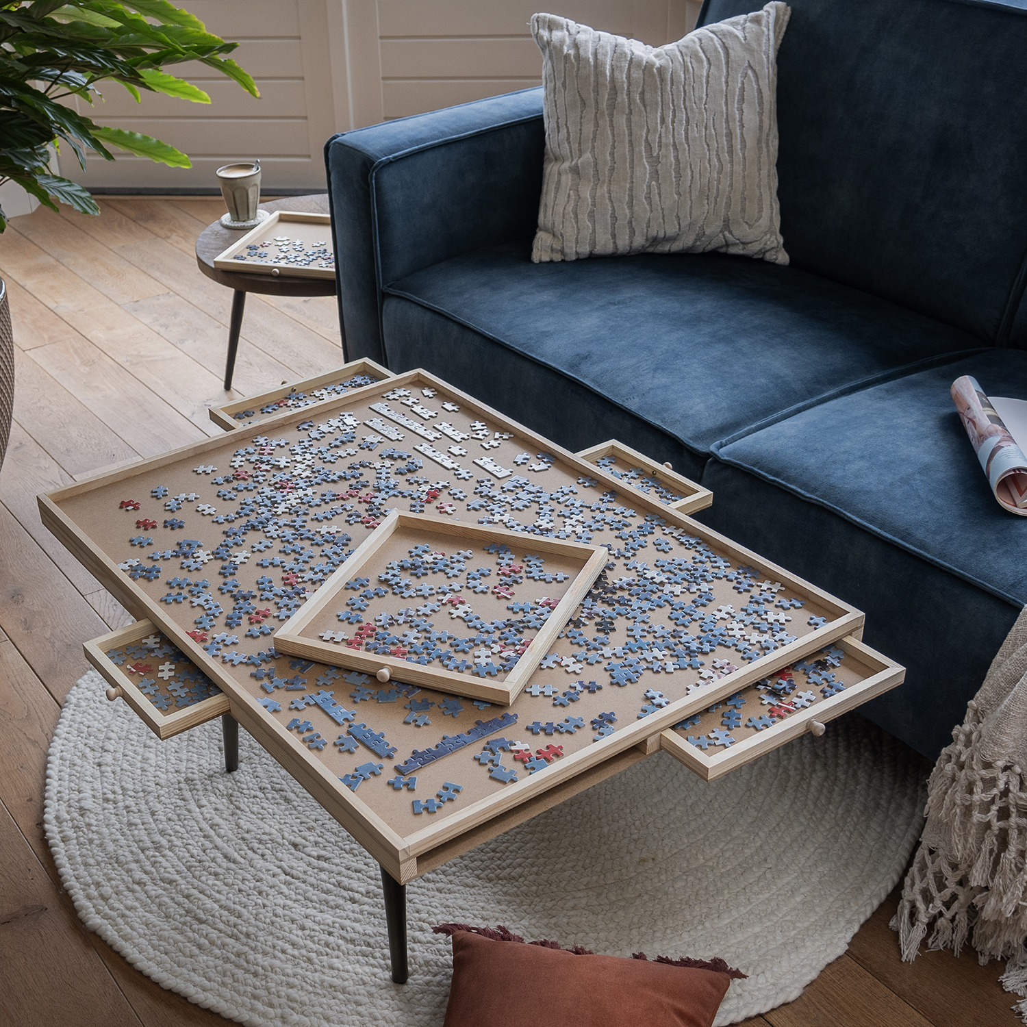 A wooden puzzle table with multiple trays is covered in jigsaw pieces. A blue sofa and a small side table are in the background.