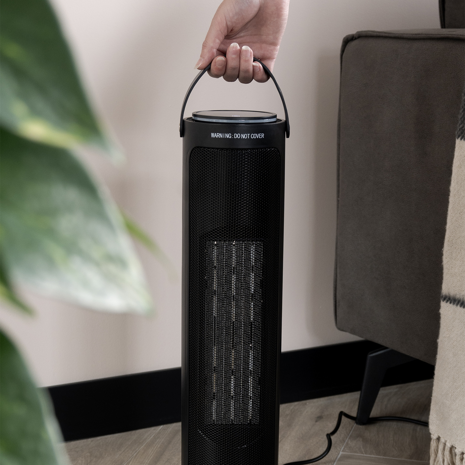 A hand holds a black heater. A grey sofa and a plant are in the background. The heater has a warning label.