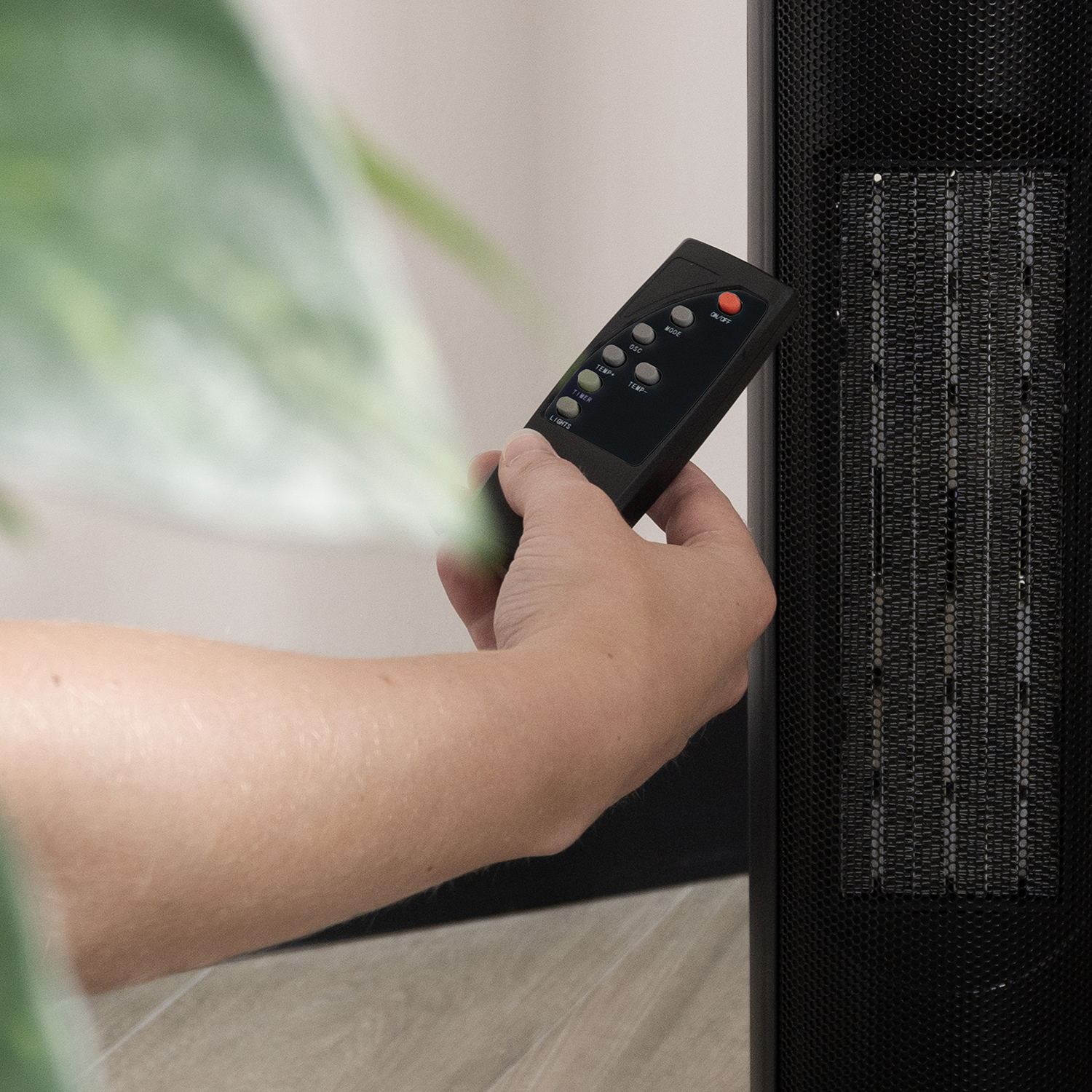 A person's hand holding a black remote control near a black heater. The remote has buttons like 'ON/OFF', 'MODE', and 'TEMP'.