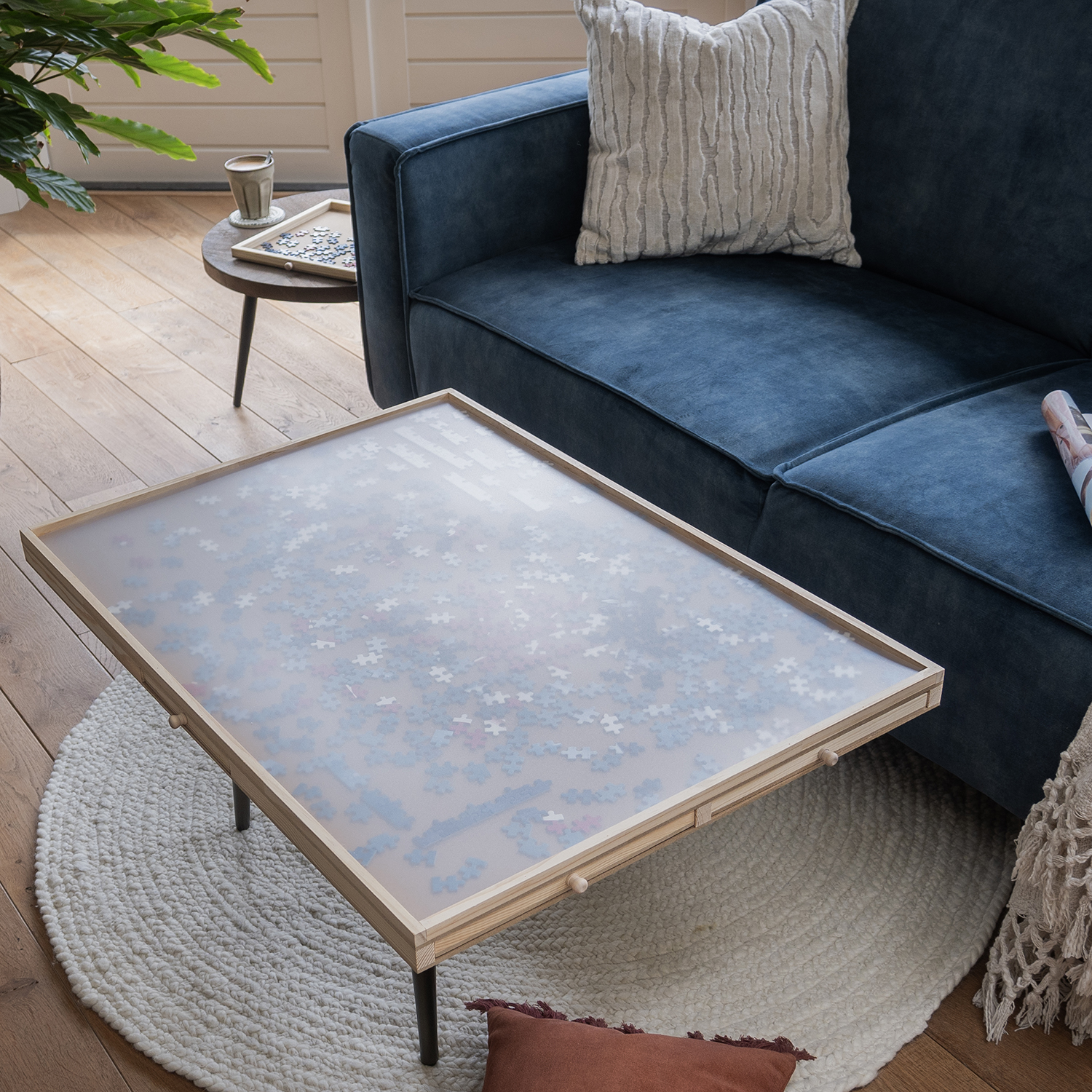 A living room scene with a coffee table holding a puzzle. A blue sofa and a small table with a cup and a puzzle tray are visible.