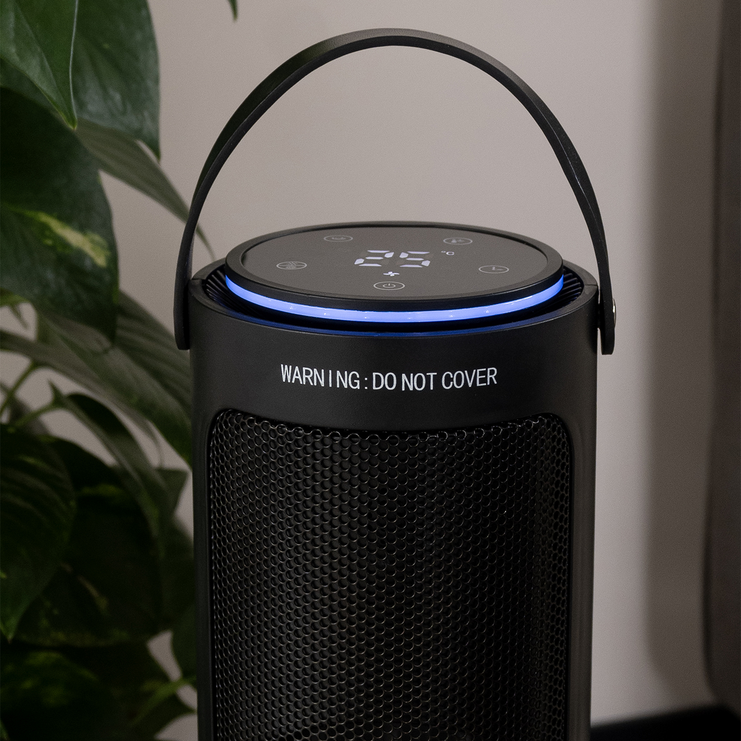 A black space heater with a handle, digital display, and warning text. Behind it is a green plant, and a light gray wall.