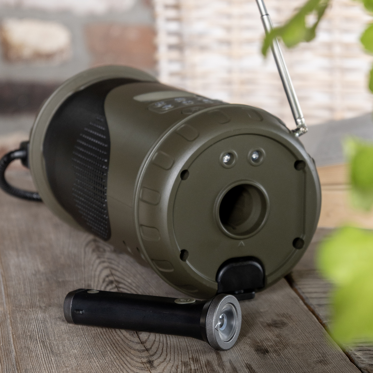 An olive-green electronic device with a black flashlight sits on a wooden surface. Brick wall background.