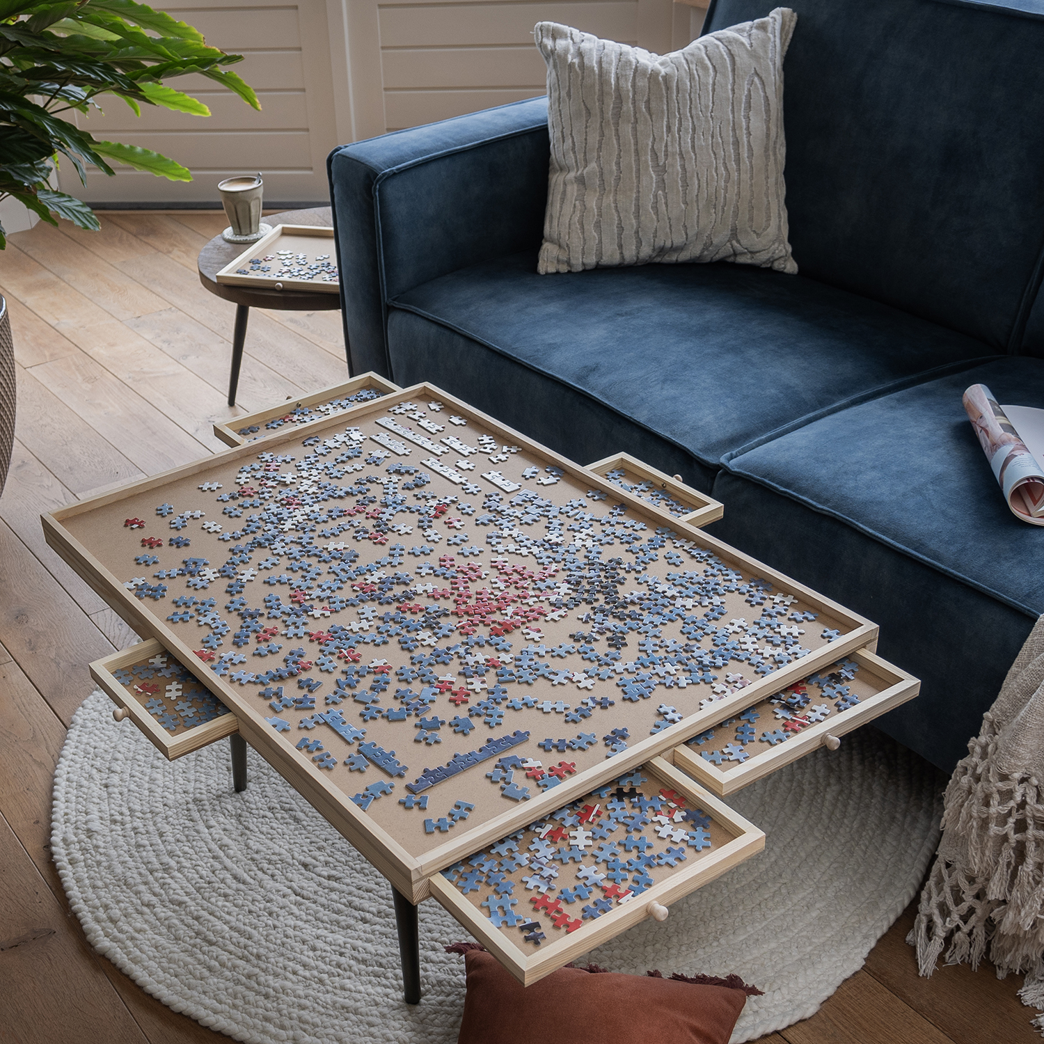 A living room with a puzzle table in the center. A blue sofa is behind it and a round rug is under the table.