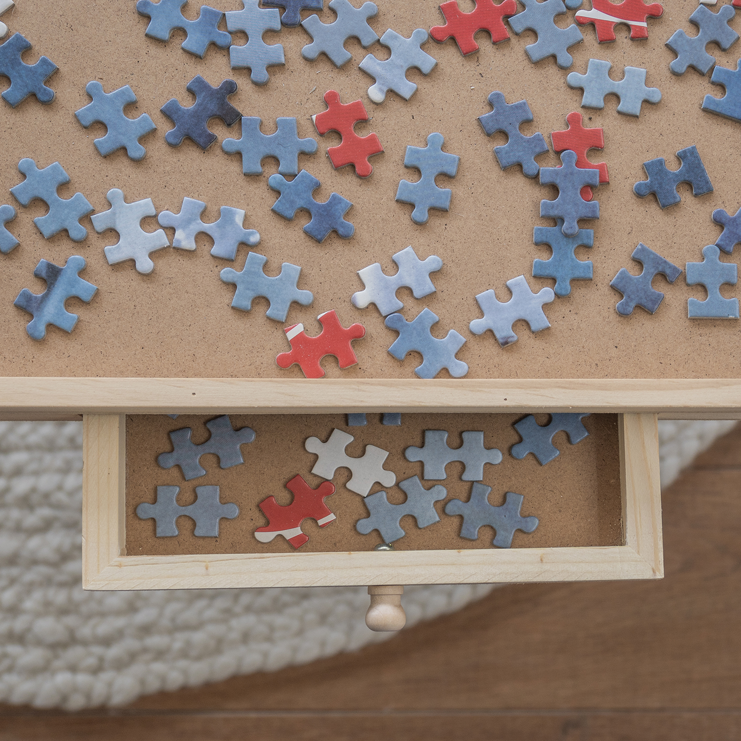 A partially completed puzzle with blue, red, and white pieces on a wooden surface. Some pieces are in a drawer.