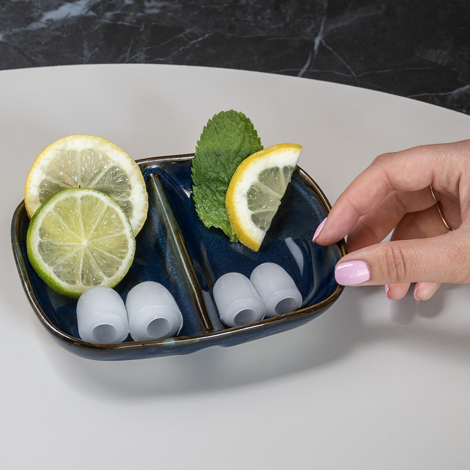 A hand holds a blue dish with lemon slices, lime slice, mint, and ice shapes. The background has a marble-like pattern.