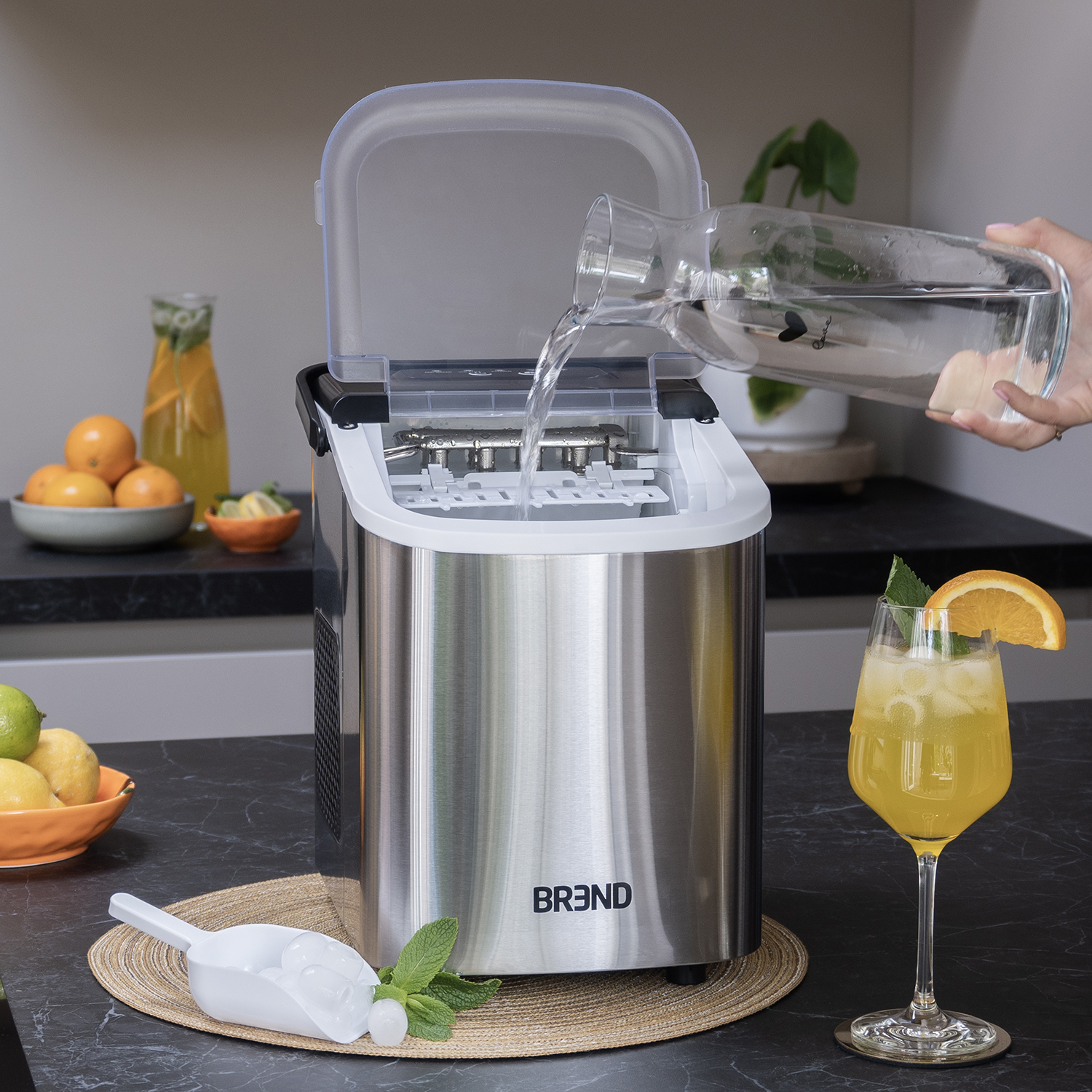 An ice maker being filled with water. A cocktail and fruits are on the counter. A scoop of ice is in front of the machine.