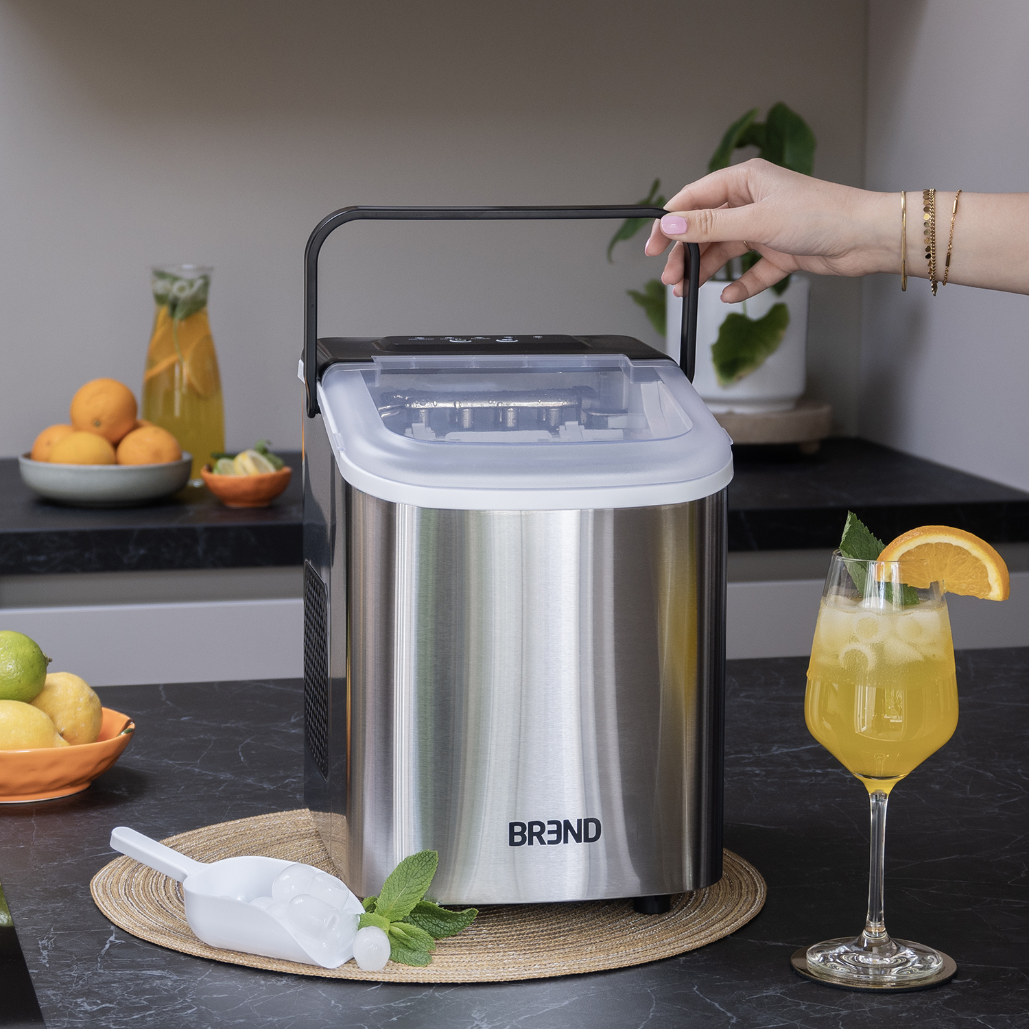 An ice machine on a counter. A hand reaches for the handle. A drink with an orange slice sits next to it, with fruit in the background.