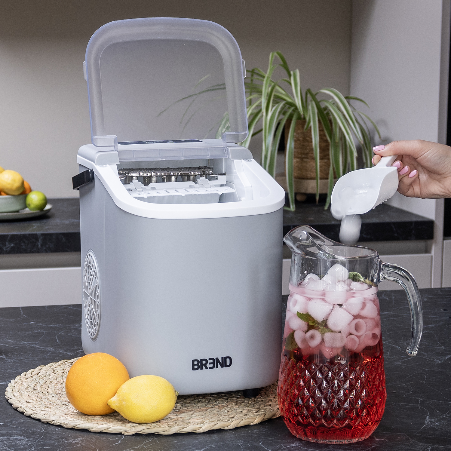 An ice maker, a pitcher of red liquid with ice and mint, fruit, and a hand scooping ice into the pitcher.