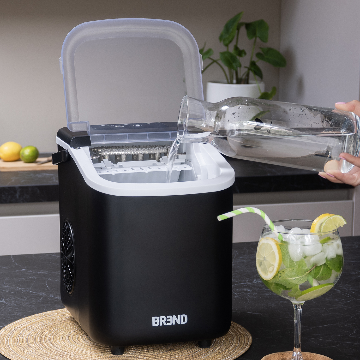 A black ice maker is being filled with water from a glass pitcher. A cocktail with lemon and mint sits nearby.