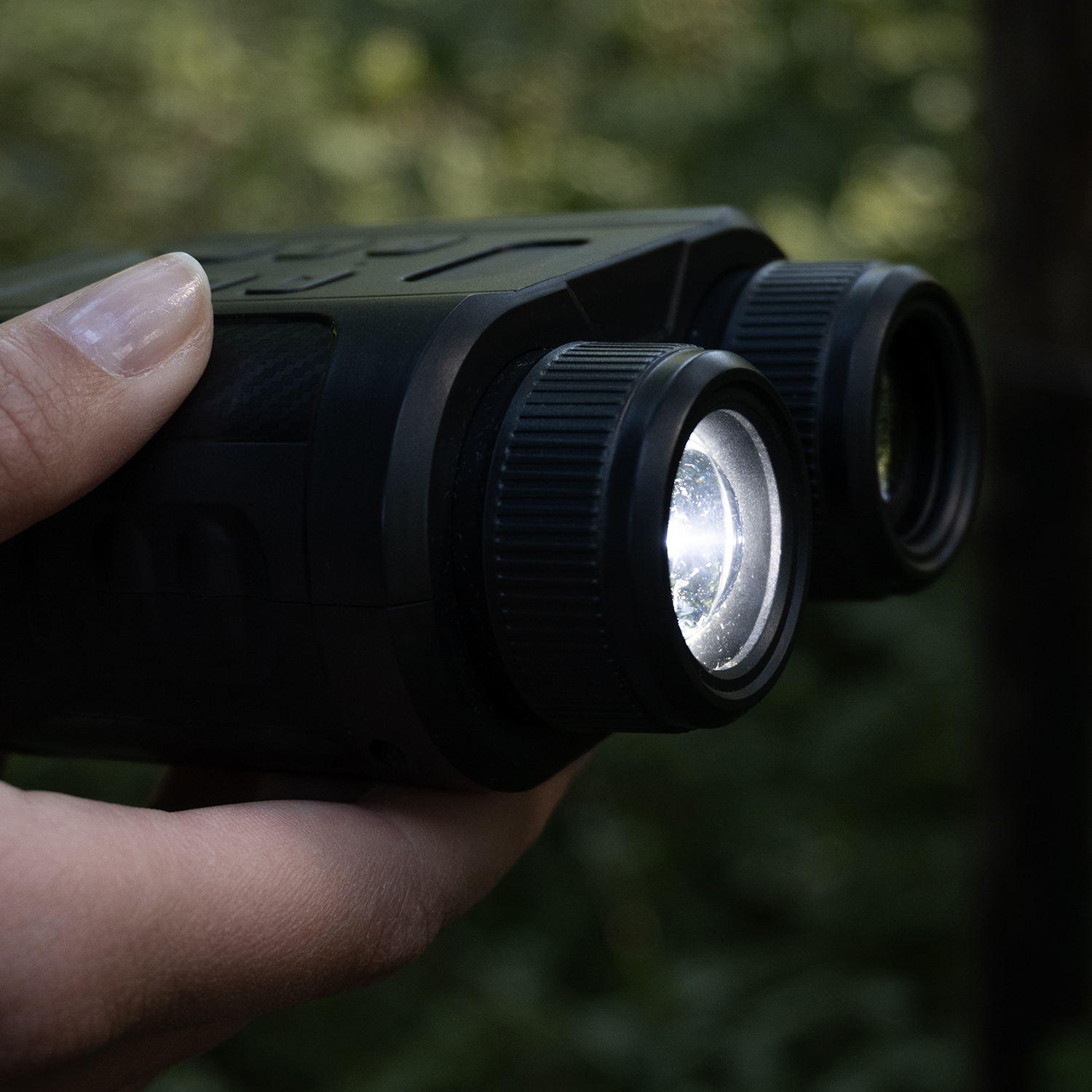 A hand holds black binoculars with a bright light in one lens. Background is out-of-focus green foliage.