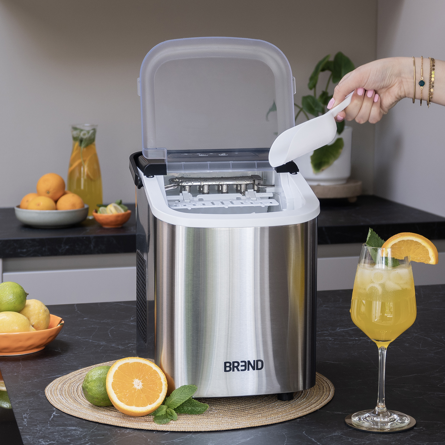 An ice maker with an open lid. A hand is scooping ice with a white scoop. A glass of cocktail with orange slice.