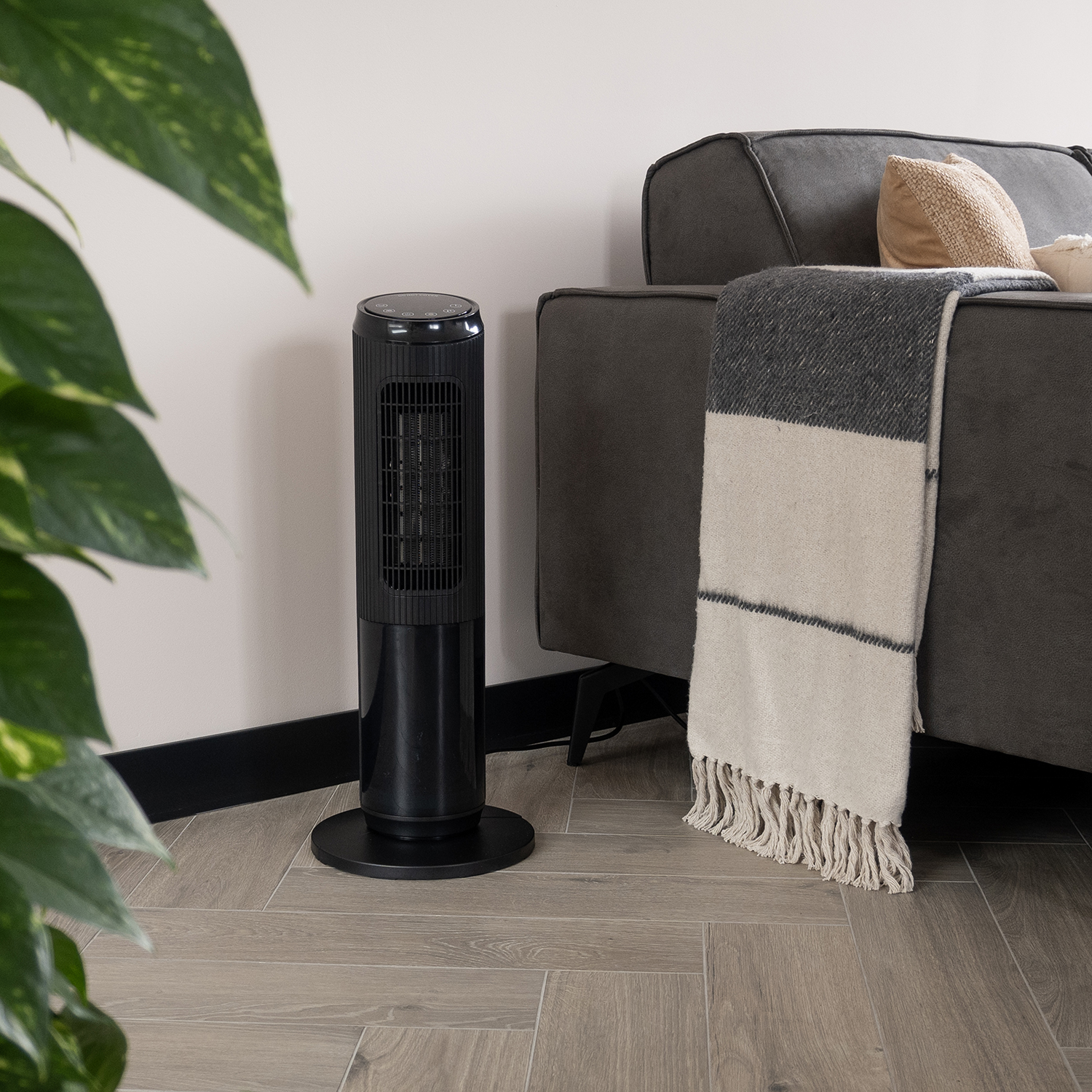 A black heater stands near a gray couch with a blanket. A plant is in the foreground. Wooden floor and cream-colored wall.