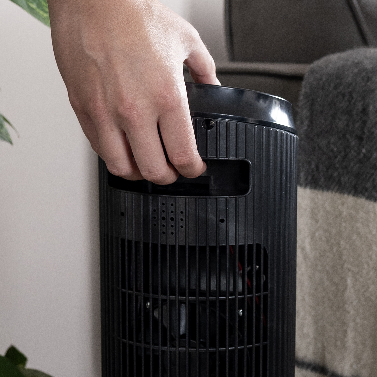 A hand holds a black fan. The fan is upright, with a grated front. A blanket and wall are in the background.