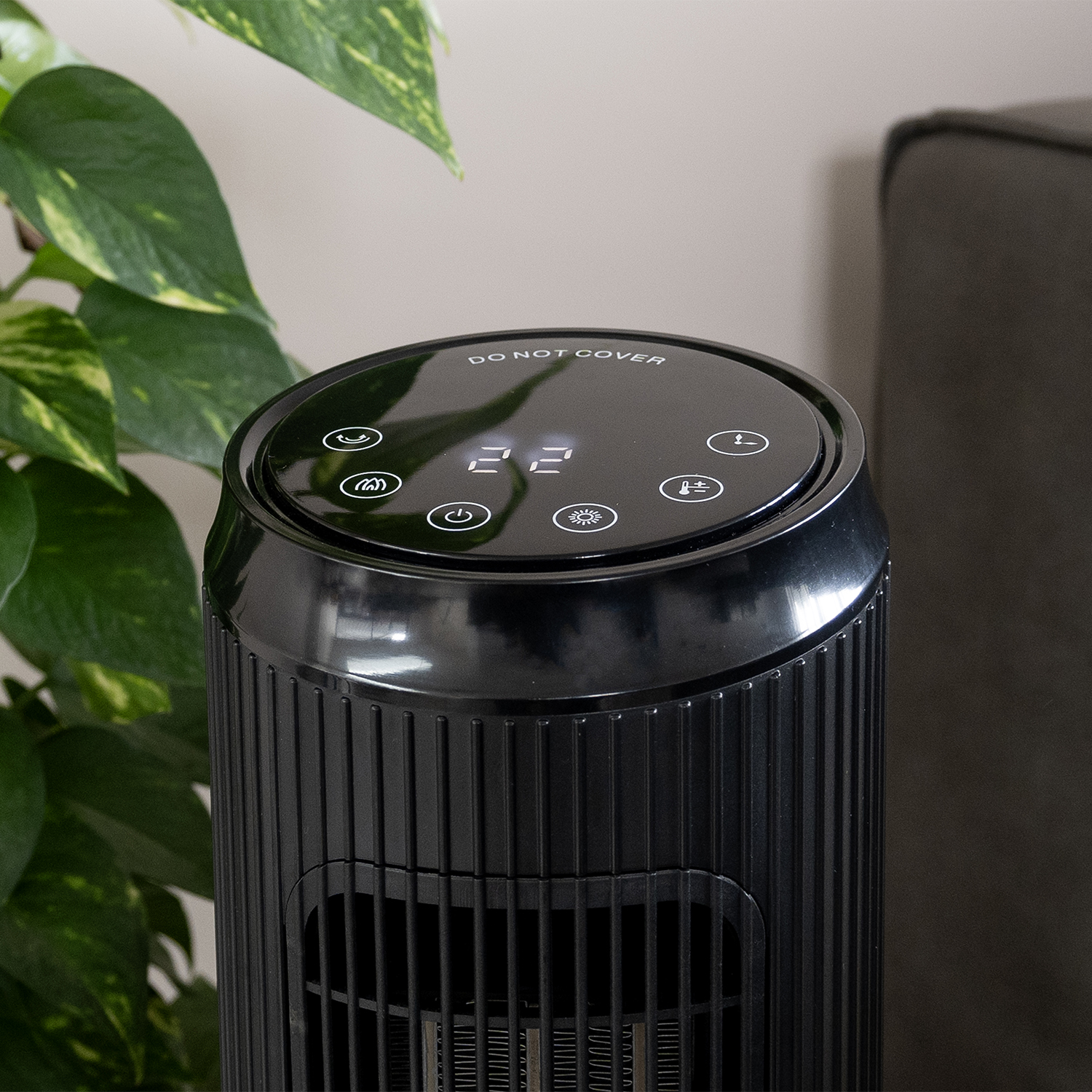 A black electric heater displays '22' on its top panel. Green leaves are to the left, and a dark gray sofa is to the right.