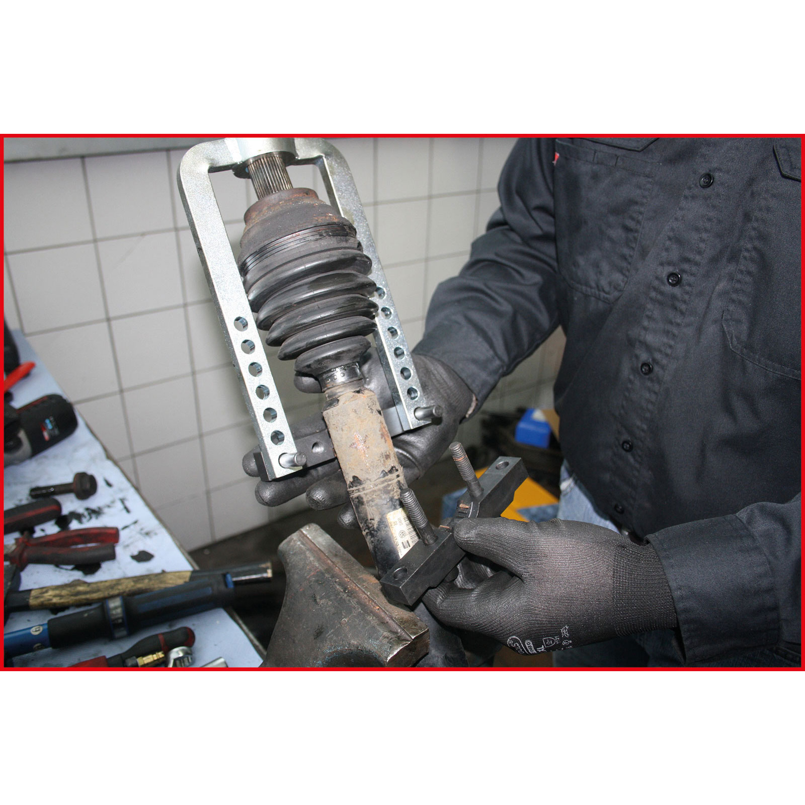 Eine Person verwendet Werkzeuge, um an einem Autoteil zu arbeiten. Sie trägt Handschuhe und ein schwarzes Hemd. Werkzeuge liegen auf einem Tisch.