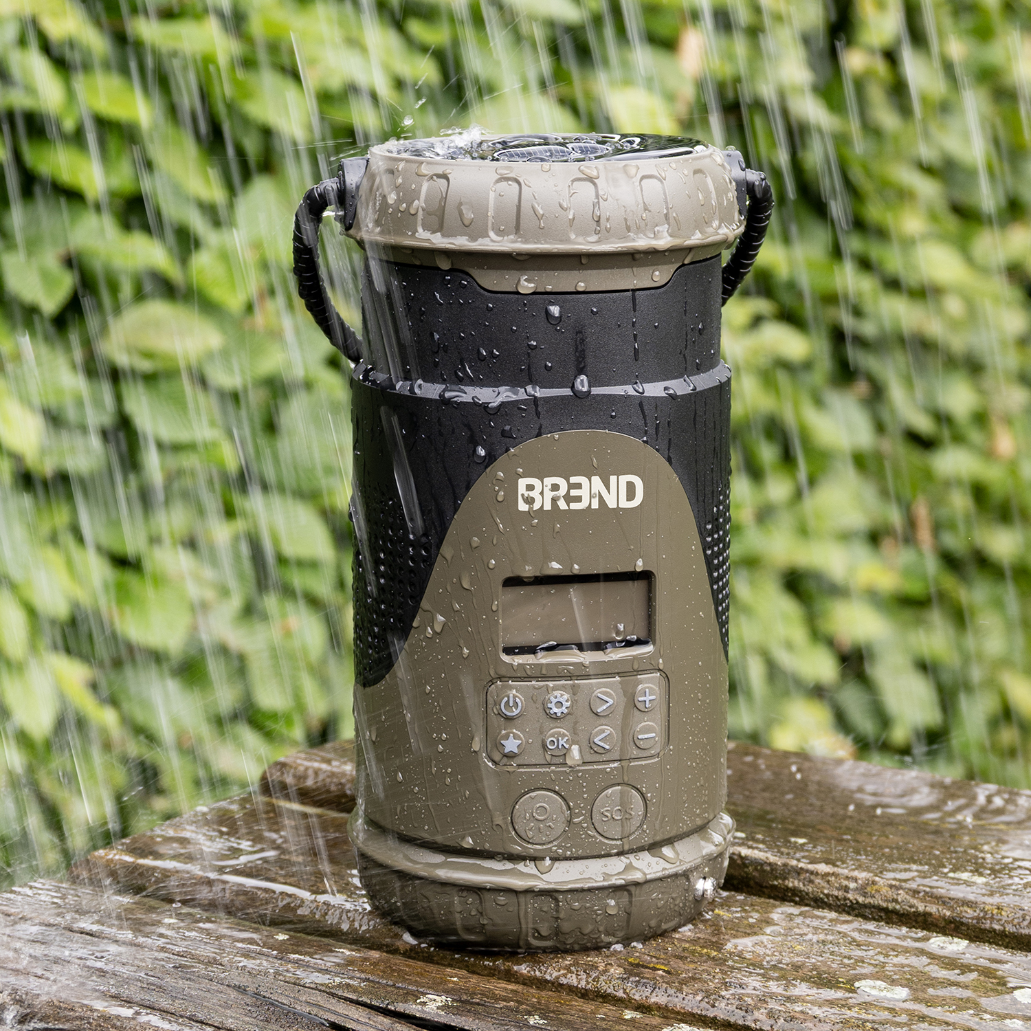 A waterproof speaker is on a wooden surface, with rain falling. The speaker is black, olive, and gray with controls and the brand name.