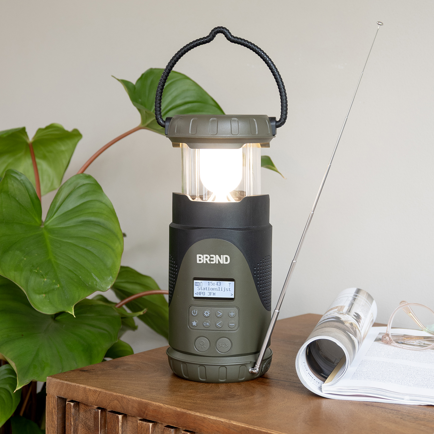 An olive green lantern with a black handle and base, a silver antenna, a magazine, and a plant on a wooden surface.