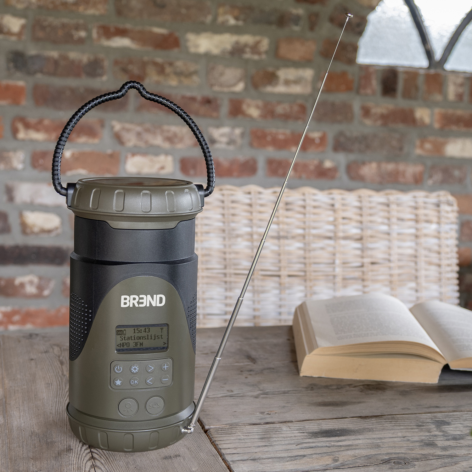 An olive-green radio with a black handle and antenna sits on a wooden table. An open book and a brick wall are in the background.