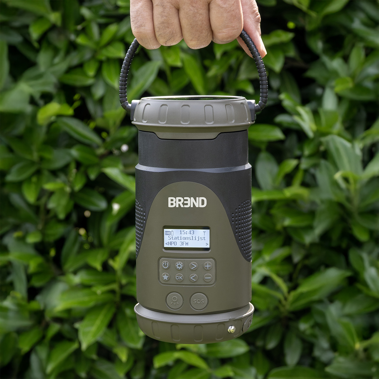 A hand holds a green and black cylindrical device with a screen. The background shows green foliage.