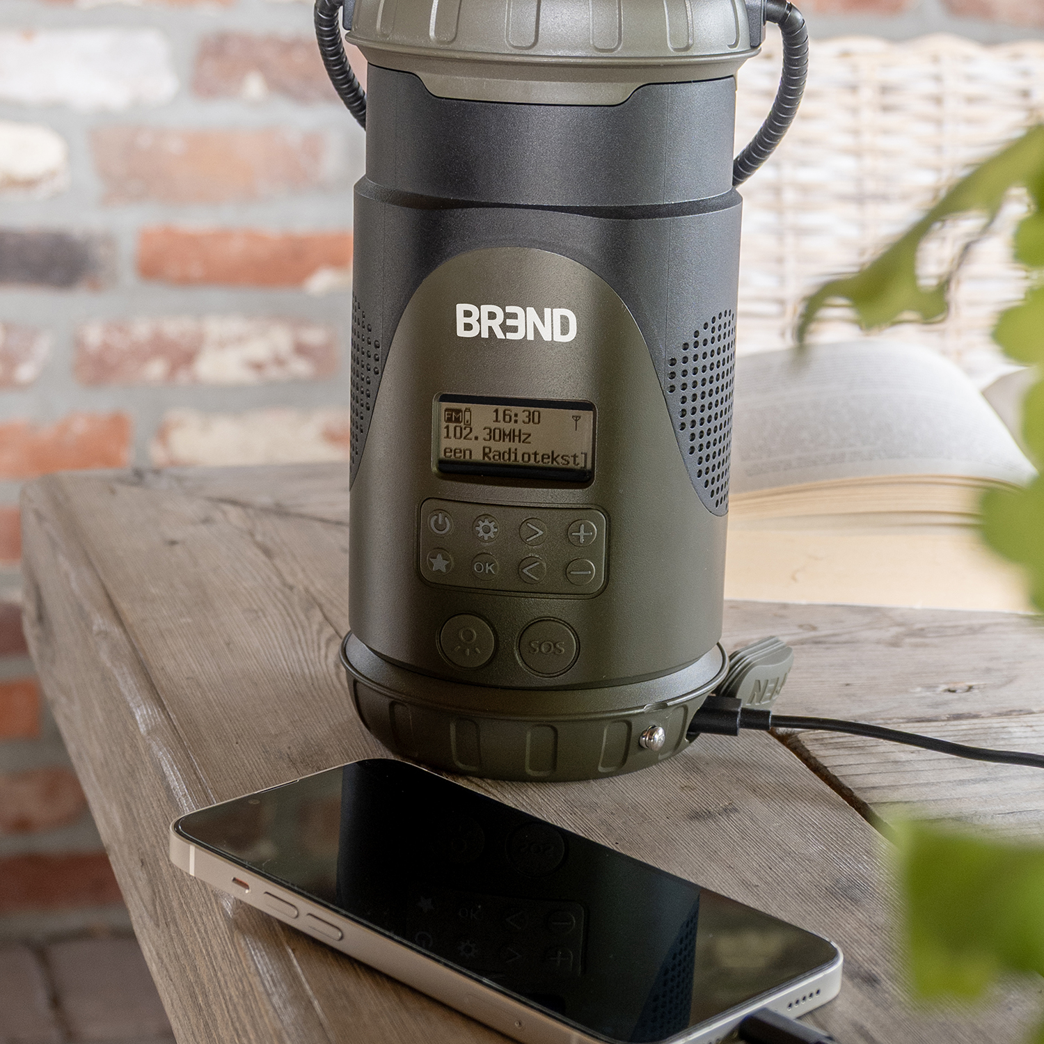 An olive green branded radio and a smartphone are on a wooden table. A cable connects the phone to the radio. A brick wall is in the background.