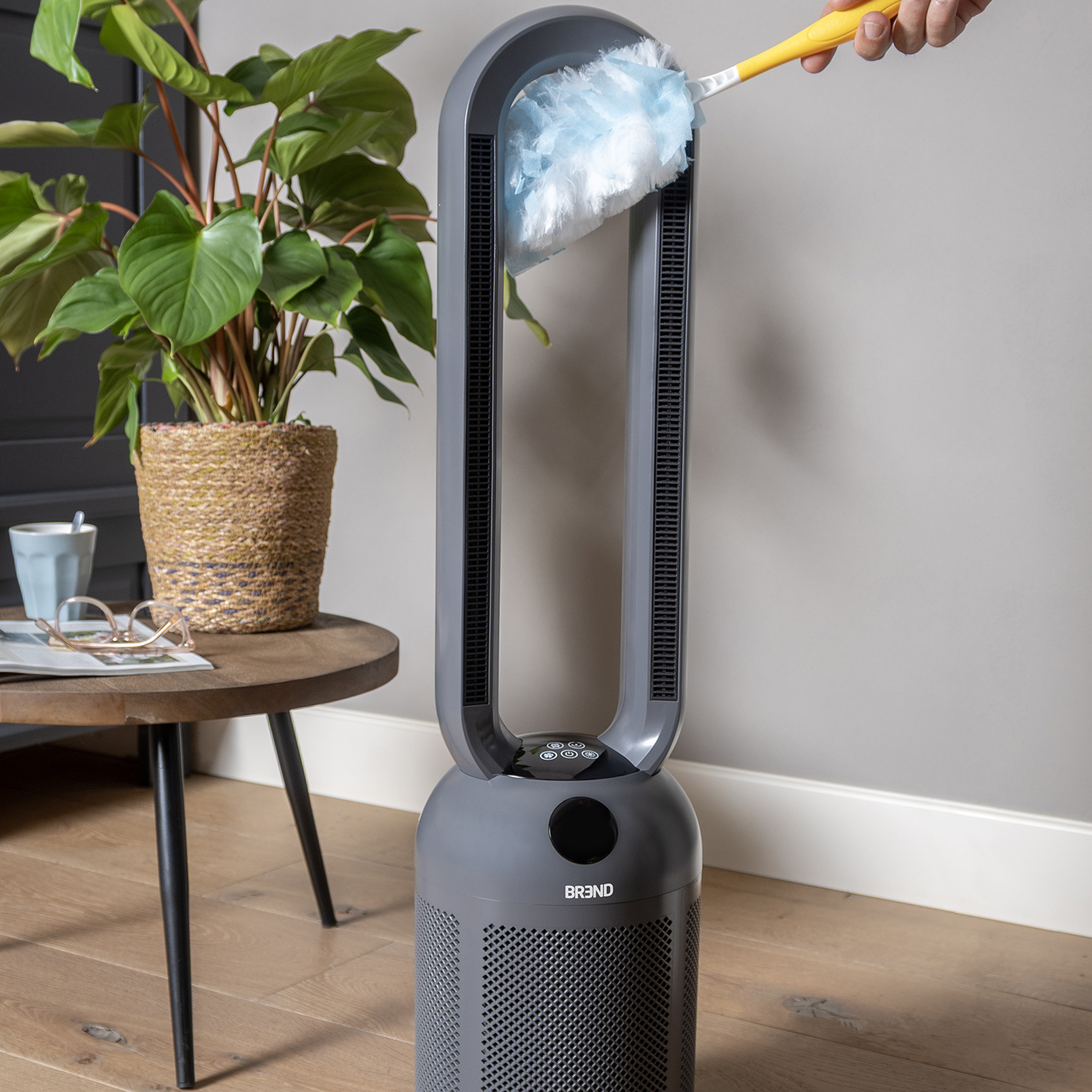 Een persoon maakt een grijze ventilator schoon met een blauwe stofdoek. Een plant in pot staat op een houten tafel naast de ventilator.