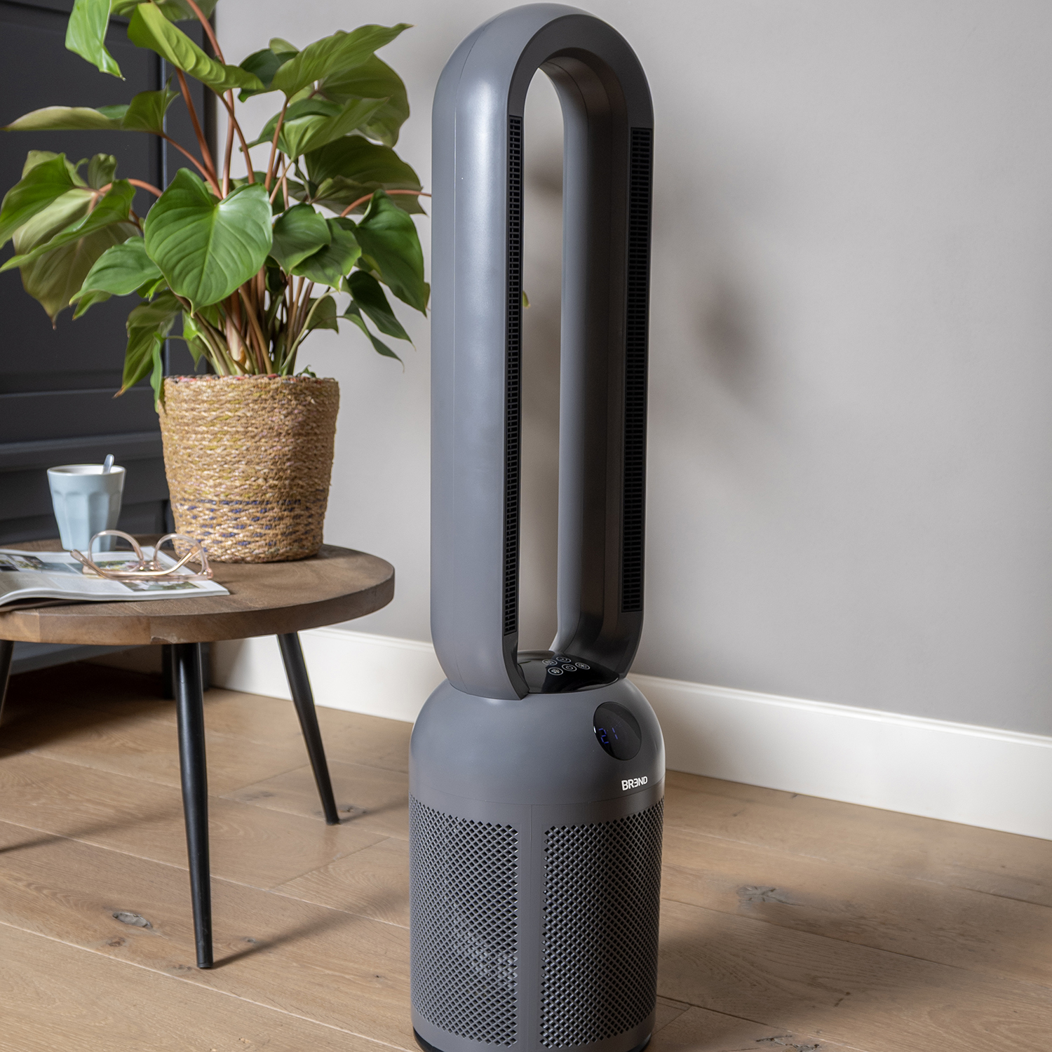 Een grijze luchtreiniger staat naast een plant in pot op een houten tafel. De achtergrond is een lichtgrijze muur en een houten vloer.