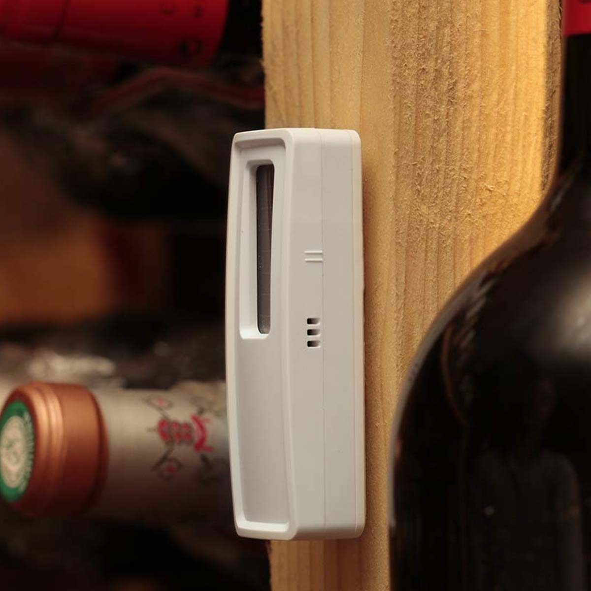 Un sensore bianco è montato su una superficie in legno, con bottiglie di vino sullo sfondo. Il sensore ha un design rettangolare con fori.