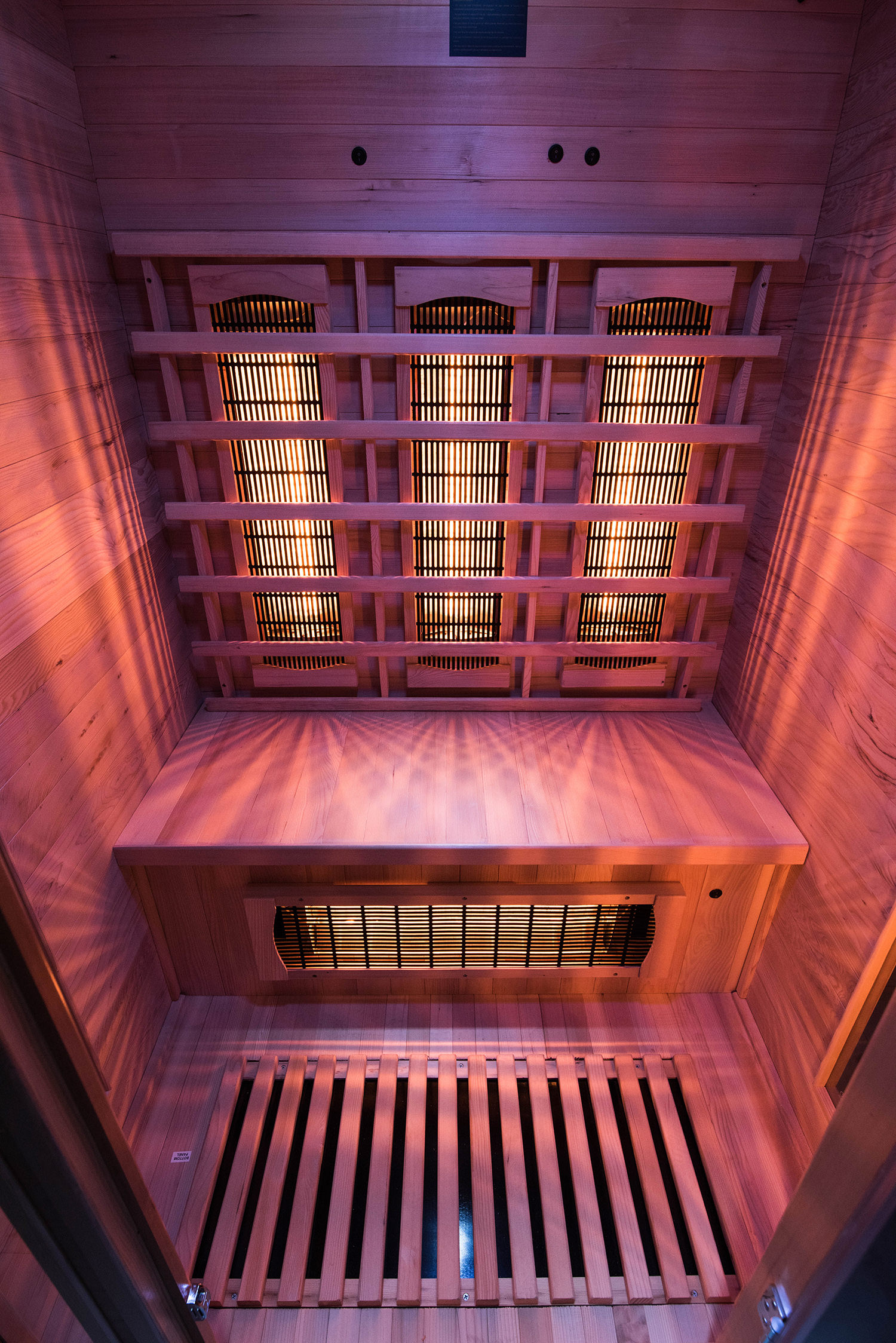Vista interior de una sauna de madera con elementos calefactores y bancos de madera. Luz cálida y rojiza ilumina.