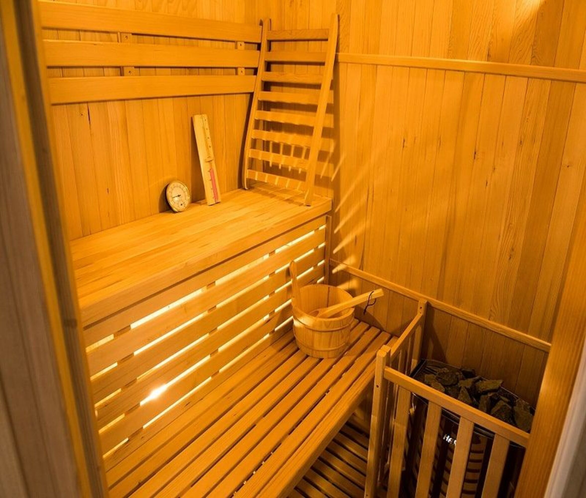 Interior de sauna de madera con bancos, una escalera, un cubo y un termómetro. Las paredes son de madera.
