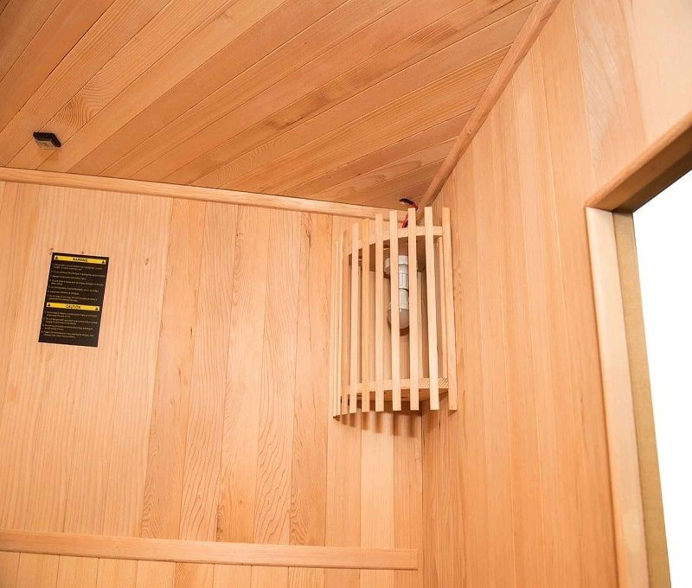 Interior de sauna de madera con una lámpara, un letrero de precaución y una puerta abierta. Paredes y techo son de madera clara.