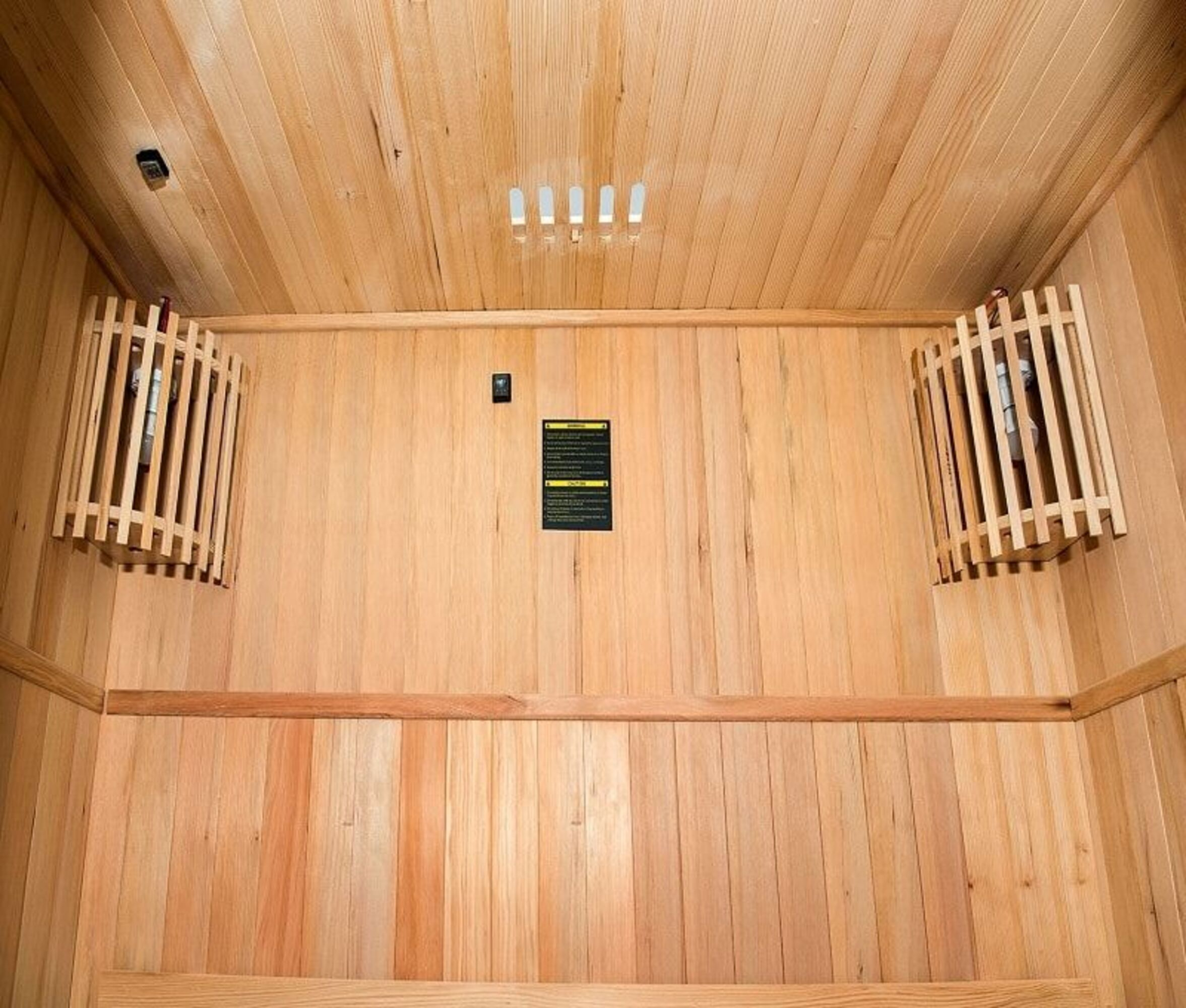 Interior de sauna de madera con bancos, paredes y techo de madera. Hay dos calentadores y un panel de control.