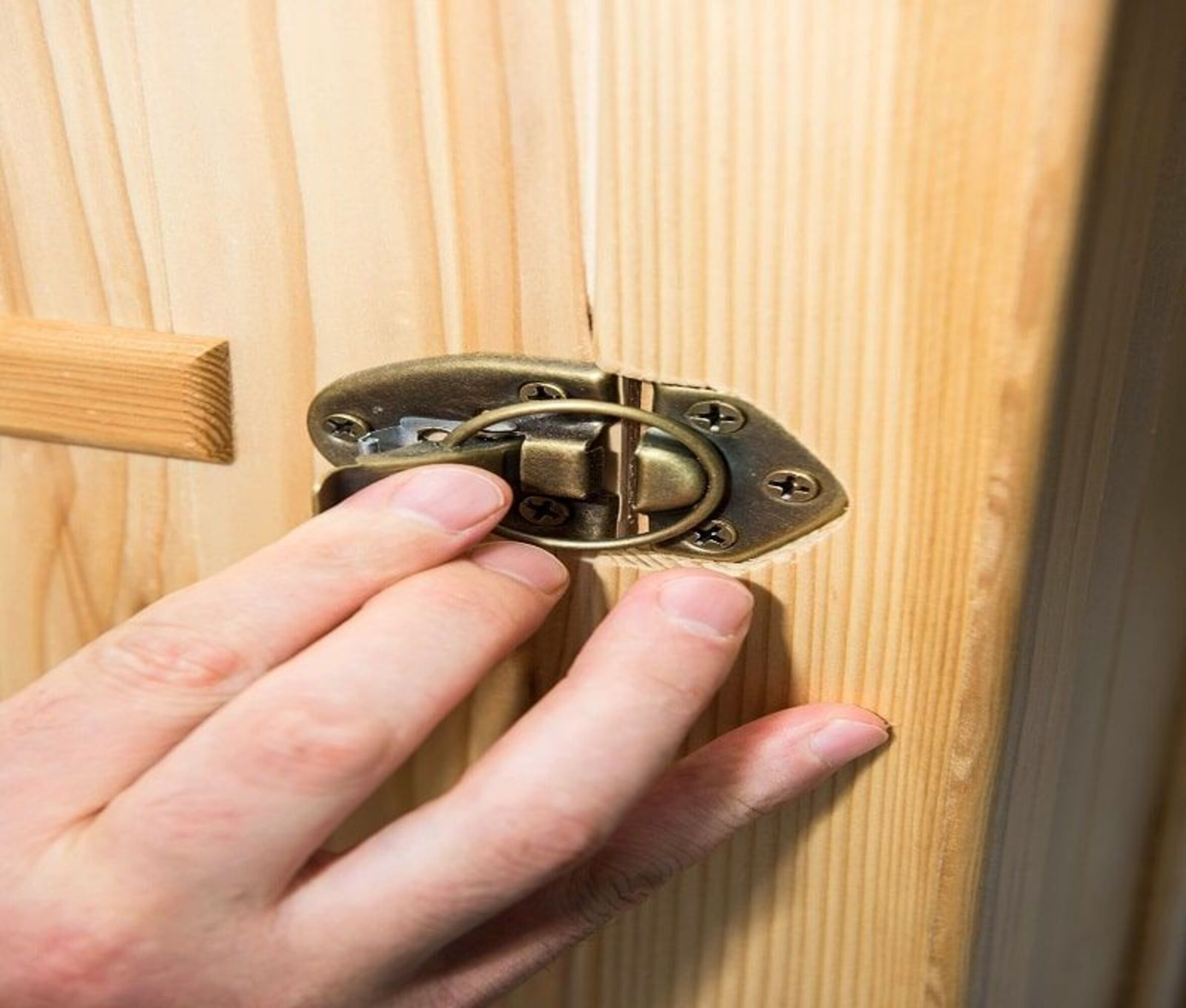 Una mano abre un pestillo de bronce en una superficie de madera. La madera es de color claro y tiene líneas verticales. El pestillo es detallado.