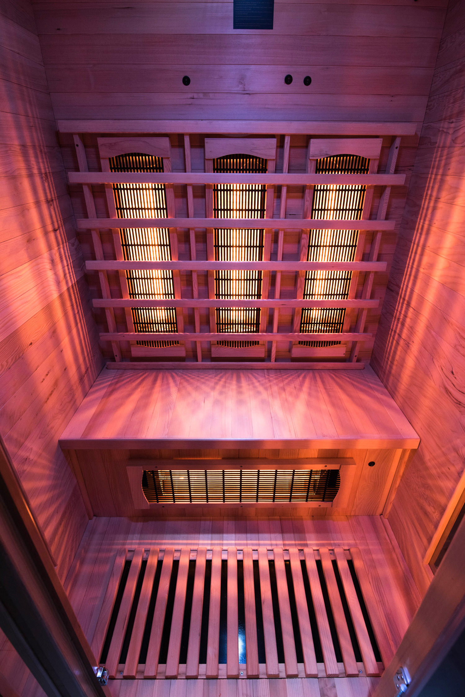 Dentro de una sauna de madera con luces naranjas. Se ven rejillas en el techo y el suelo. Las paredes son de madera.