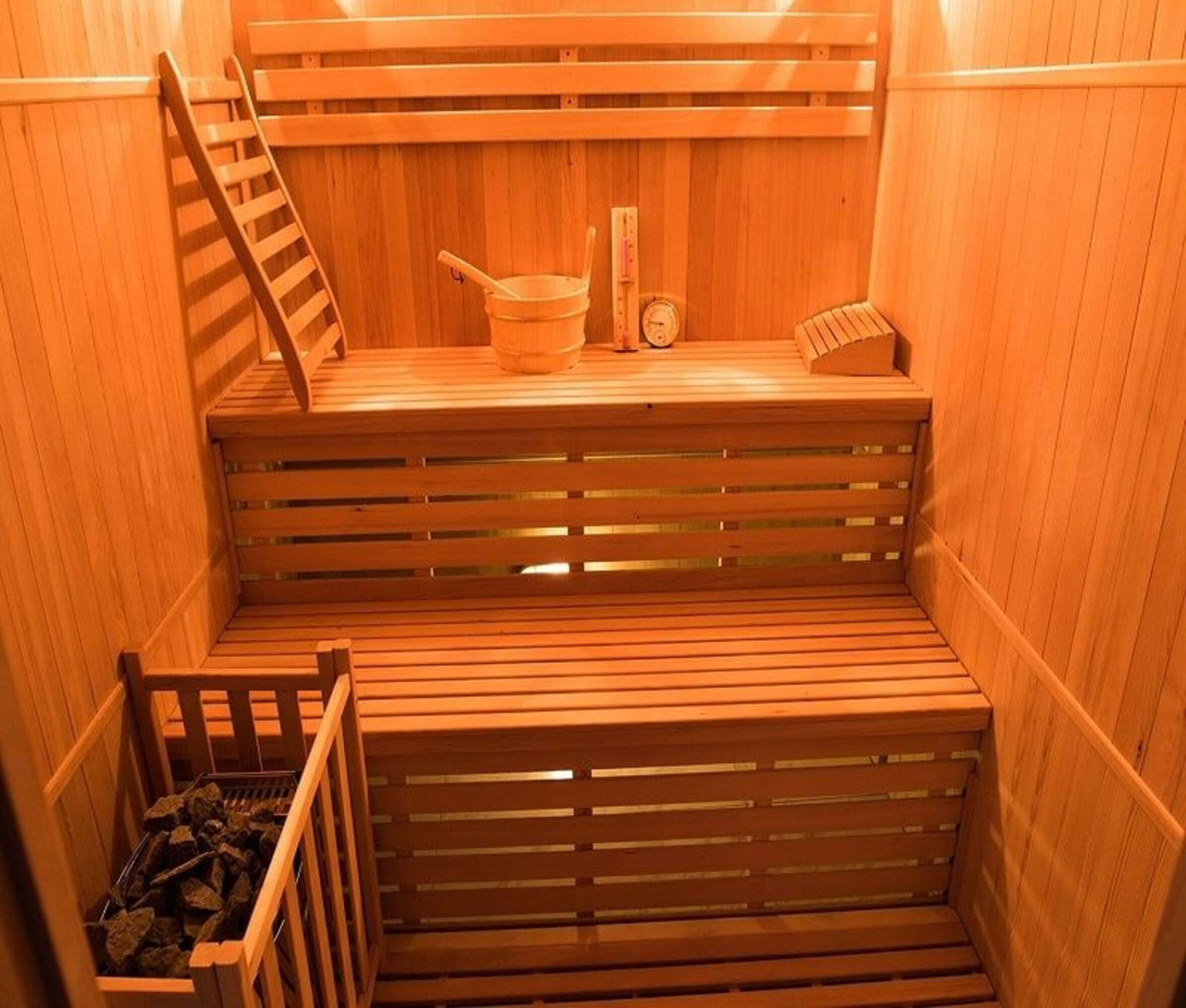 Interior de sauna de madera con escalera, bancos, cubo, piedras y termómetro.
