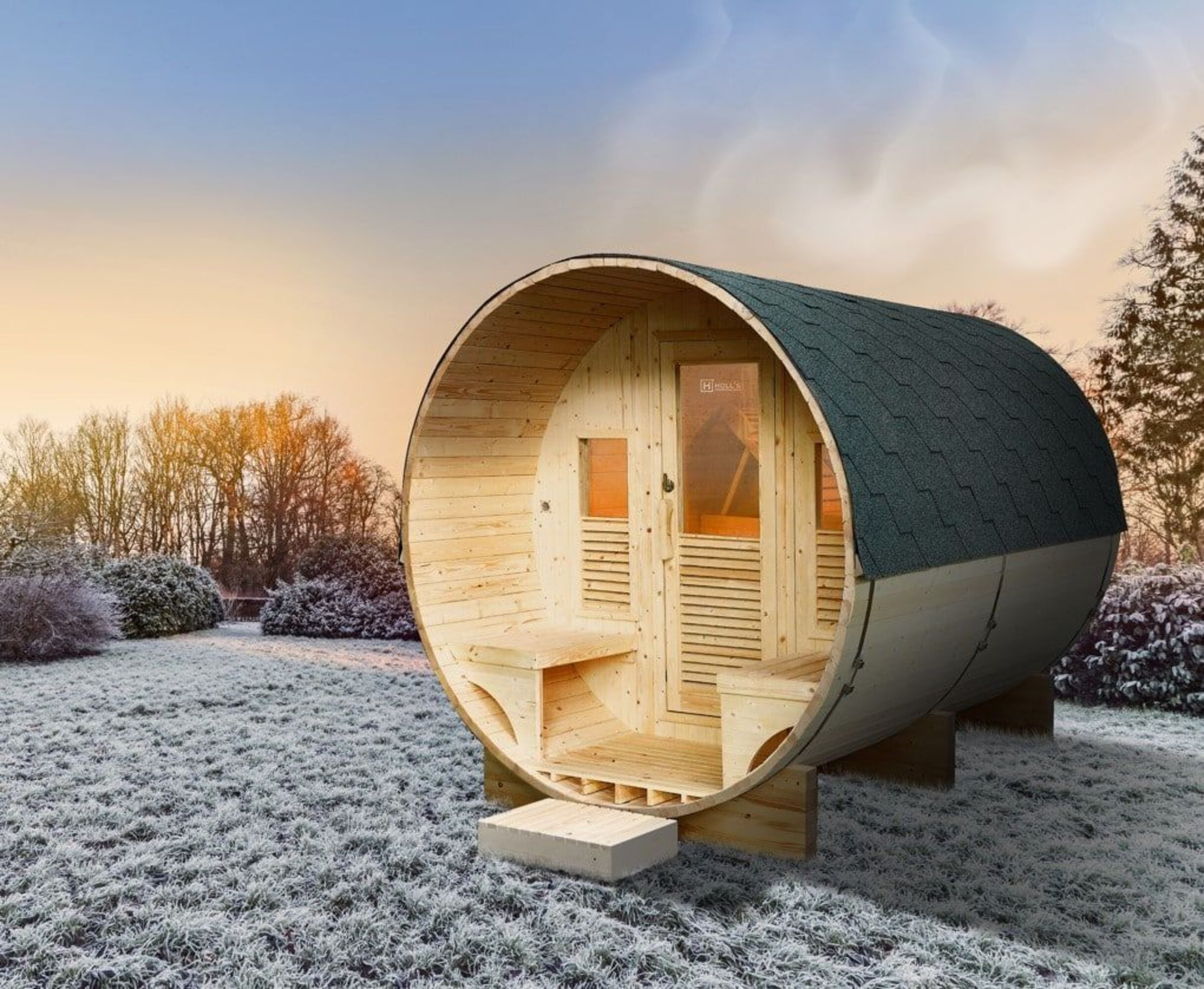 Sauna de barril de madera en césped escarchado. Tiene techo verde, ventanas y una entrada.