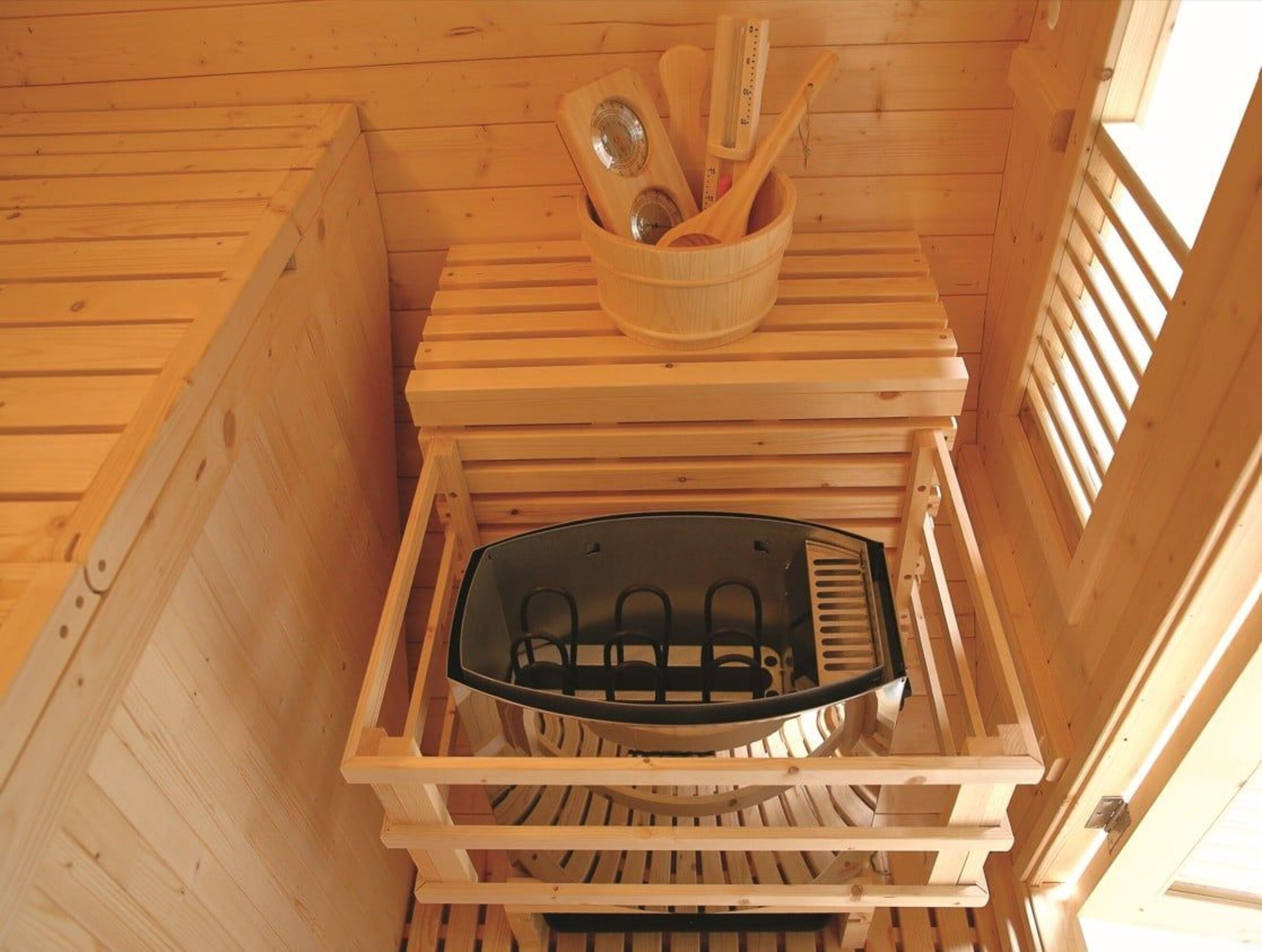 Interior de sauna de madera con calentador, cubo con herramientas y banco de listones. Paredes y techo de madera.