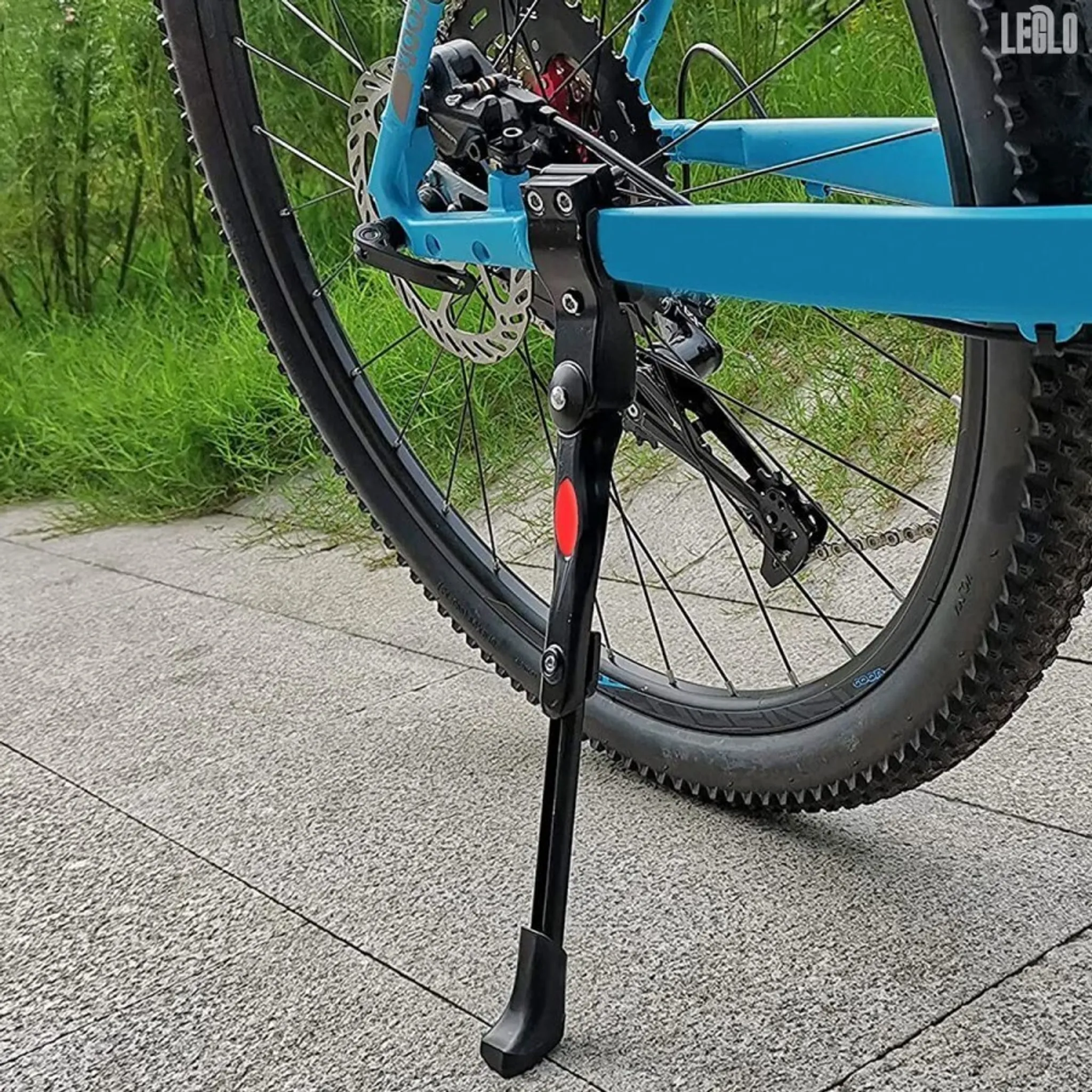 Ein blaues Fahrrad mit einem schwarzen Ständer. Das Fahrrad steht auf grauem Pflaster, im Hintergrund ist Gras zu sehen.