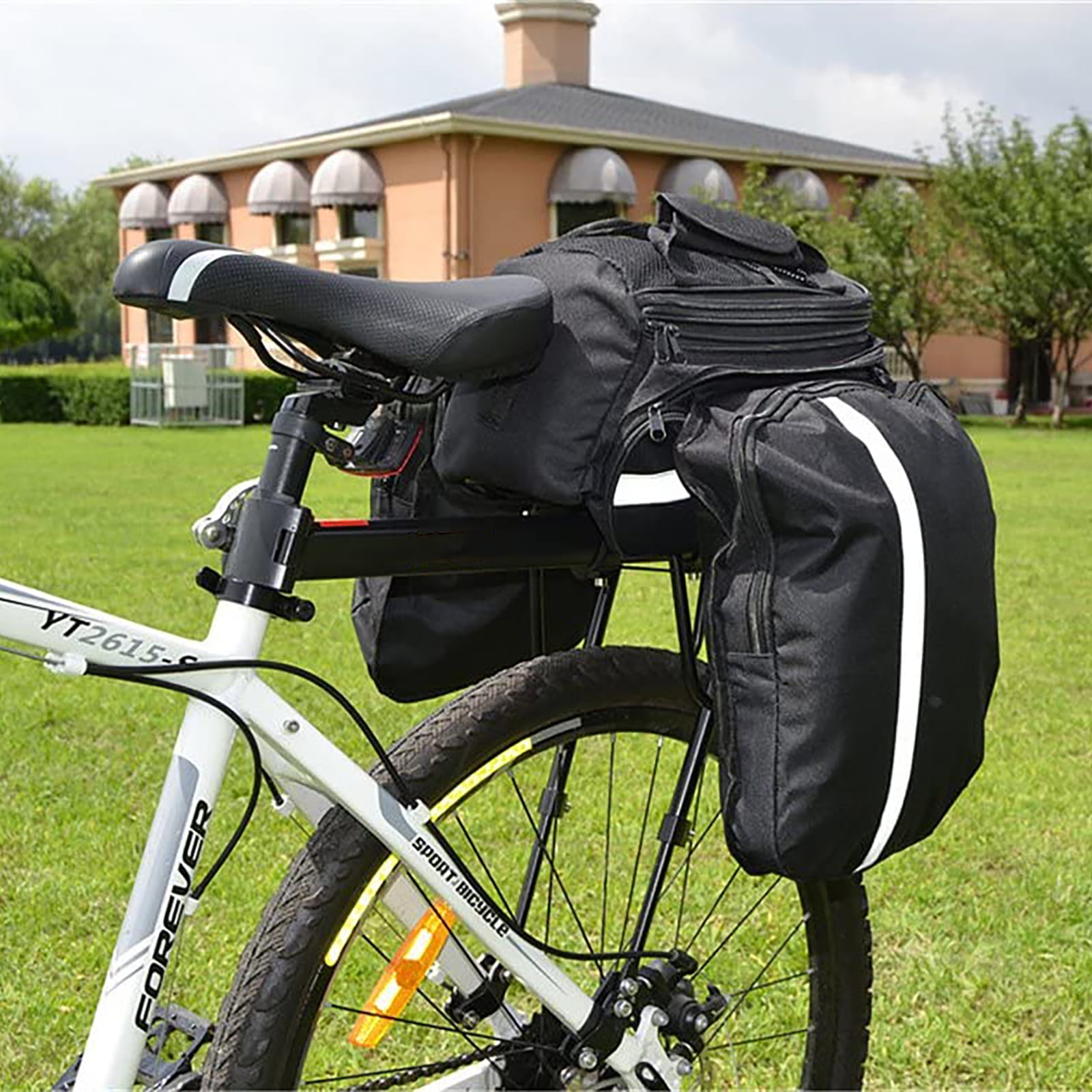 Ein Fahrrad mit schwarzen Packtaschen und weißem Rahmen. Im Hintergrund ein Haus mit Markisen auf einer grünen Wiese.