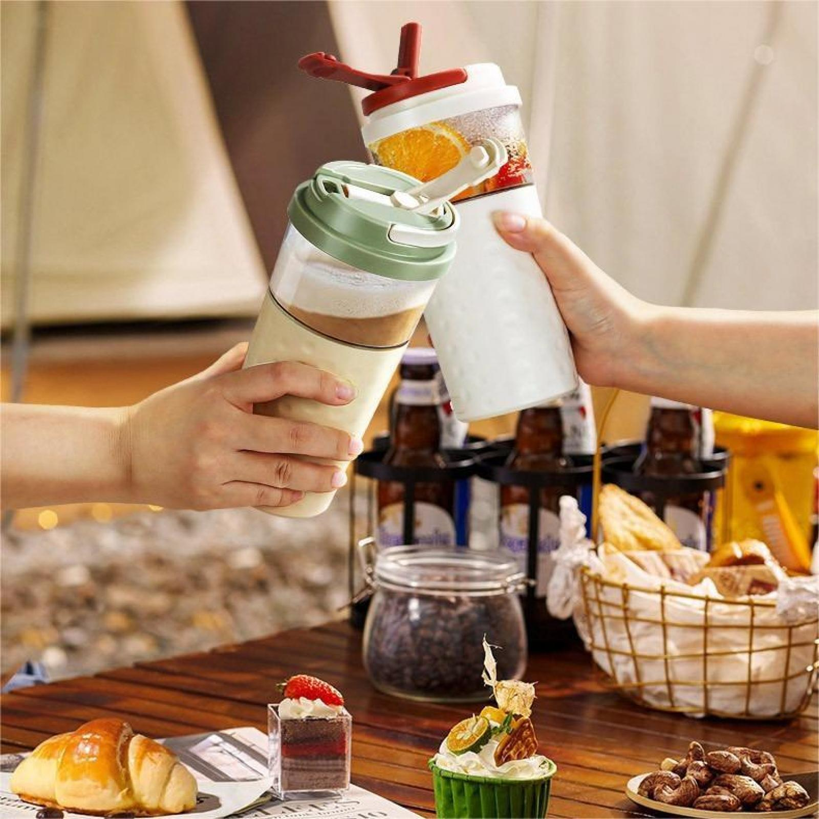 Hände halten zwei Mixer mit Getränken, Essen und Snacks auf einem Holztisch. Flaschen und ein Korb sind im Hintergrund.