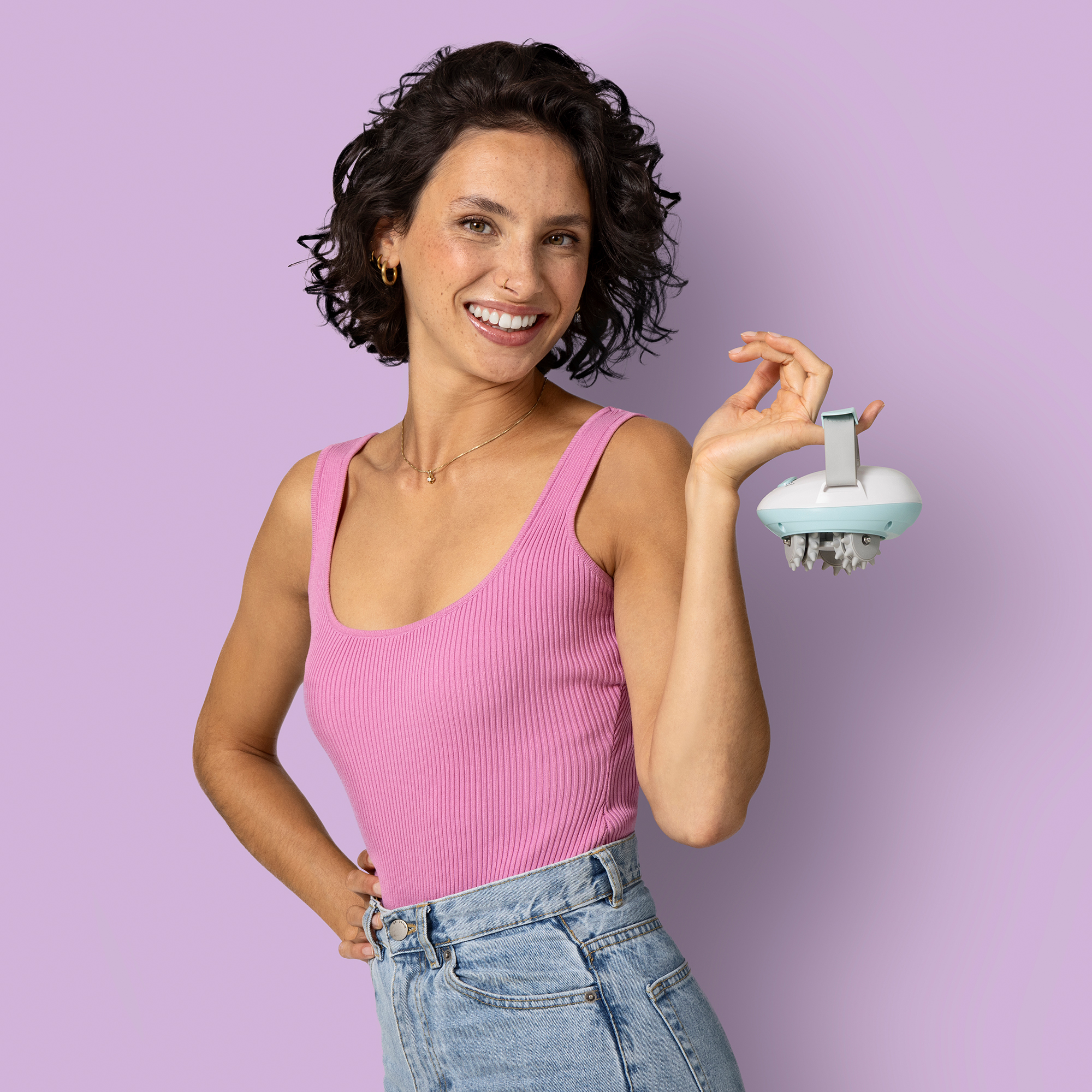Kobieta w różowym topie trzyma urządzenie. Uśmiecha się do kamery, ma ciemne kręcone włosy i niebieskie dżinsy. Tło jest fioletowe.
