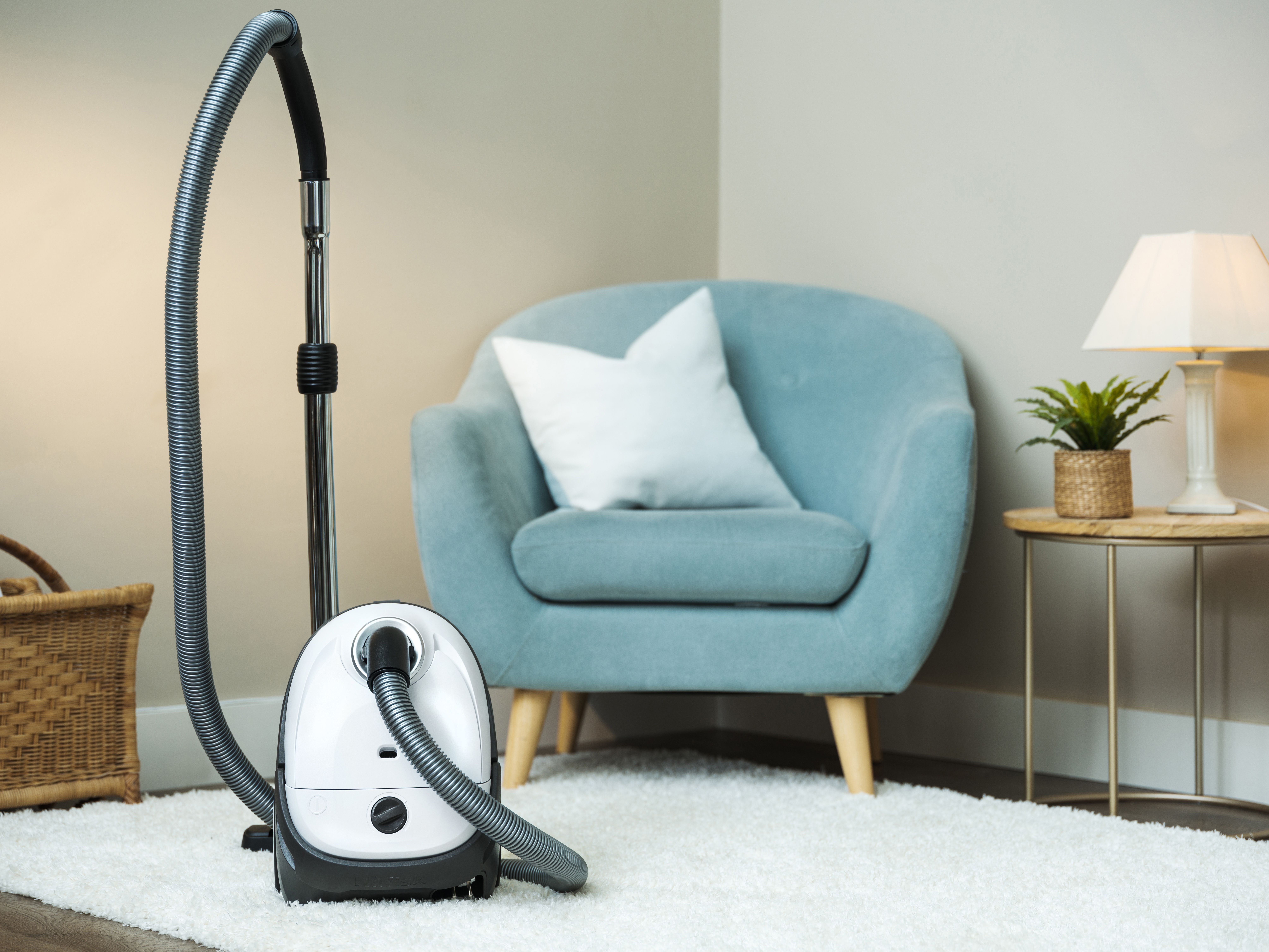 Odkurzacz stoi na białym dywanie. Niebieski fotel z białą poduszką w tle.