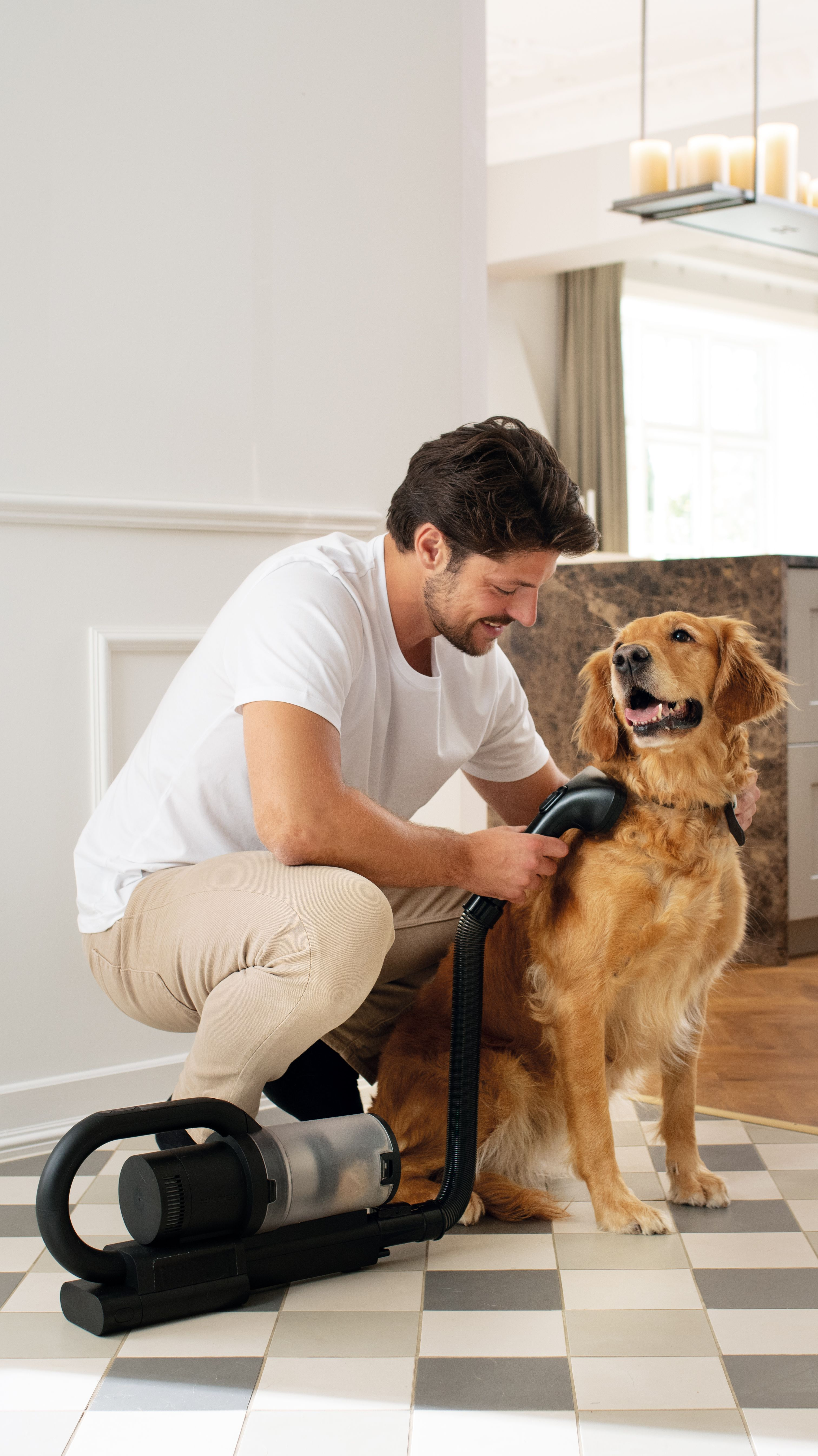 Un homme brosse un golden retriever avec un aspirateur. Le chien est assis. La pièce a des planchers de tuiles et des murs blancs.