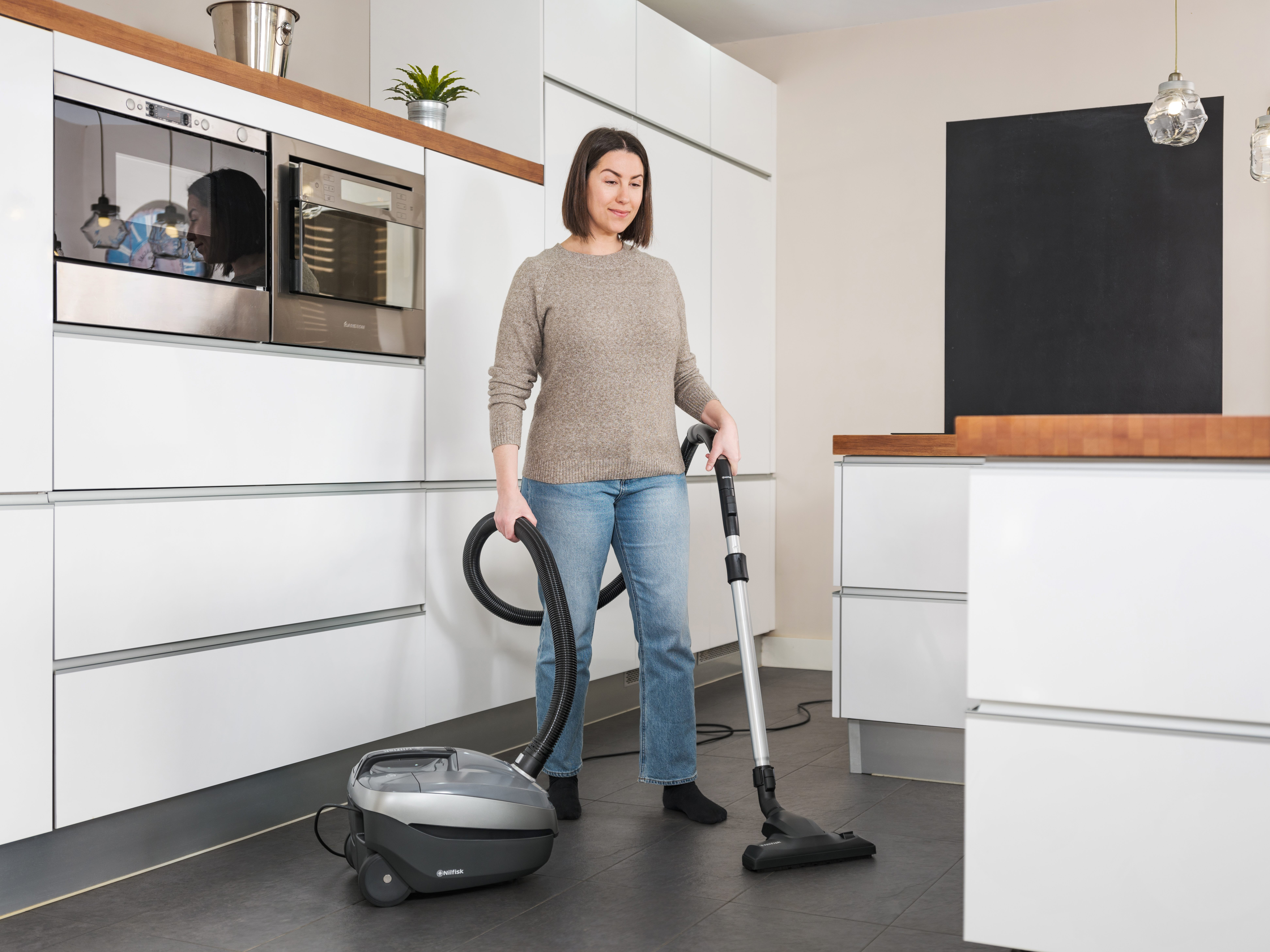 Mujer aspira un piso de cocina. Lleva jeans y un suéter. La cocina tiene gabinetes y electrodomésticos blancos.