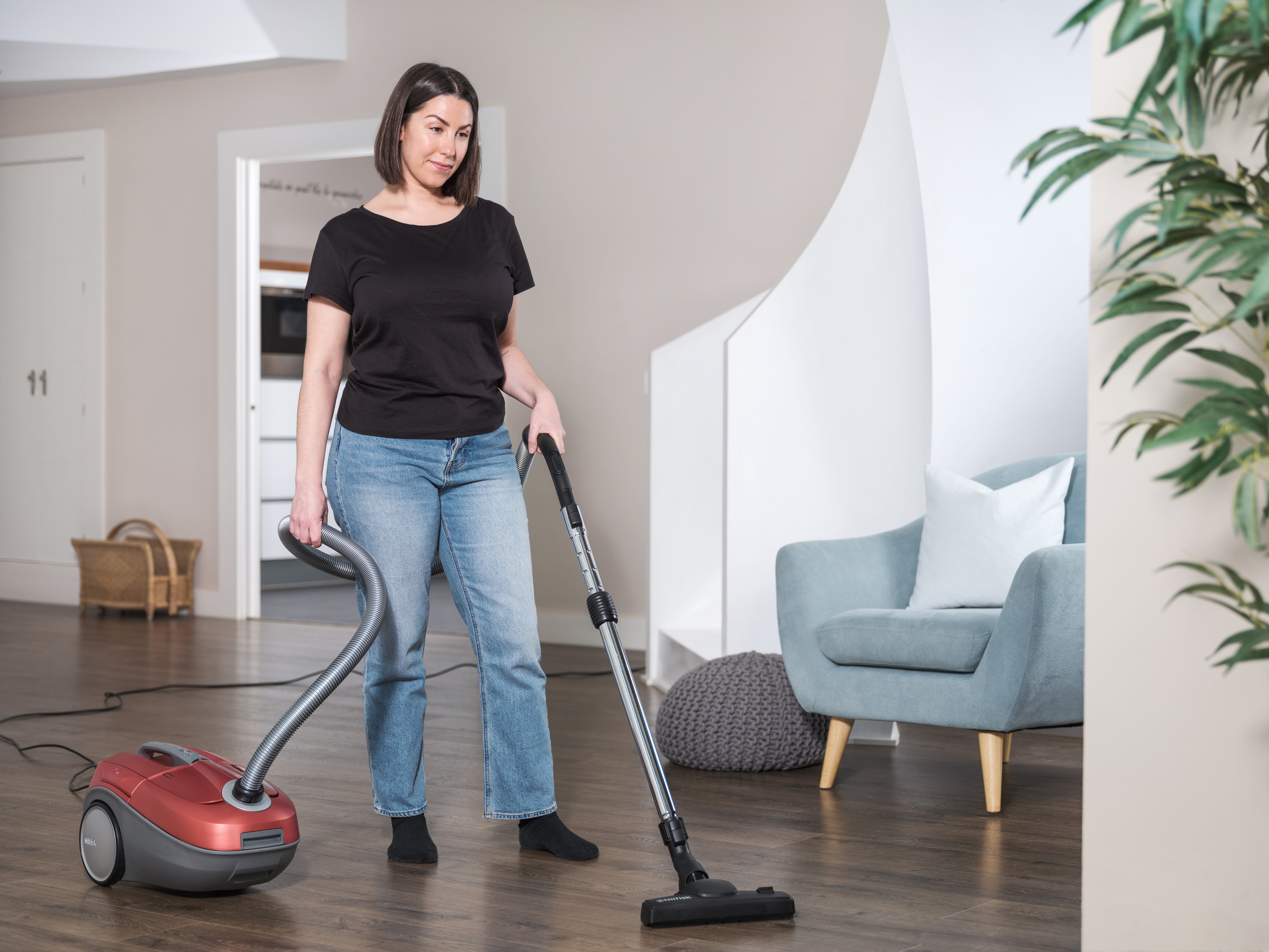 Kobieta odkurza drewnianą podłogę. Ma na sobie czarną koszulę i dżinsy. Po lewej stronie czerwony odkurzacz.