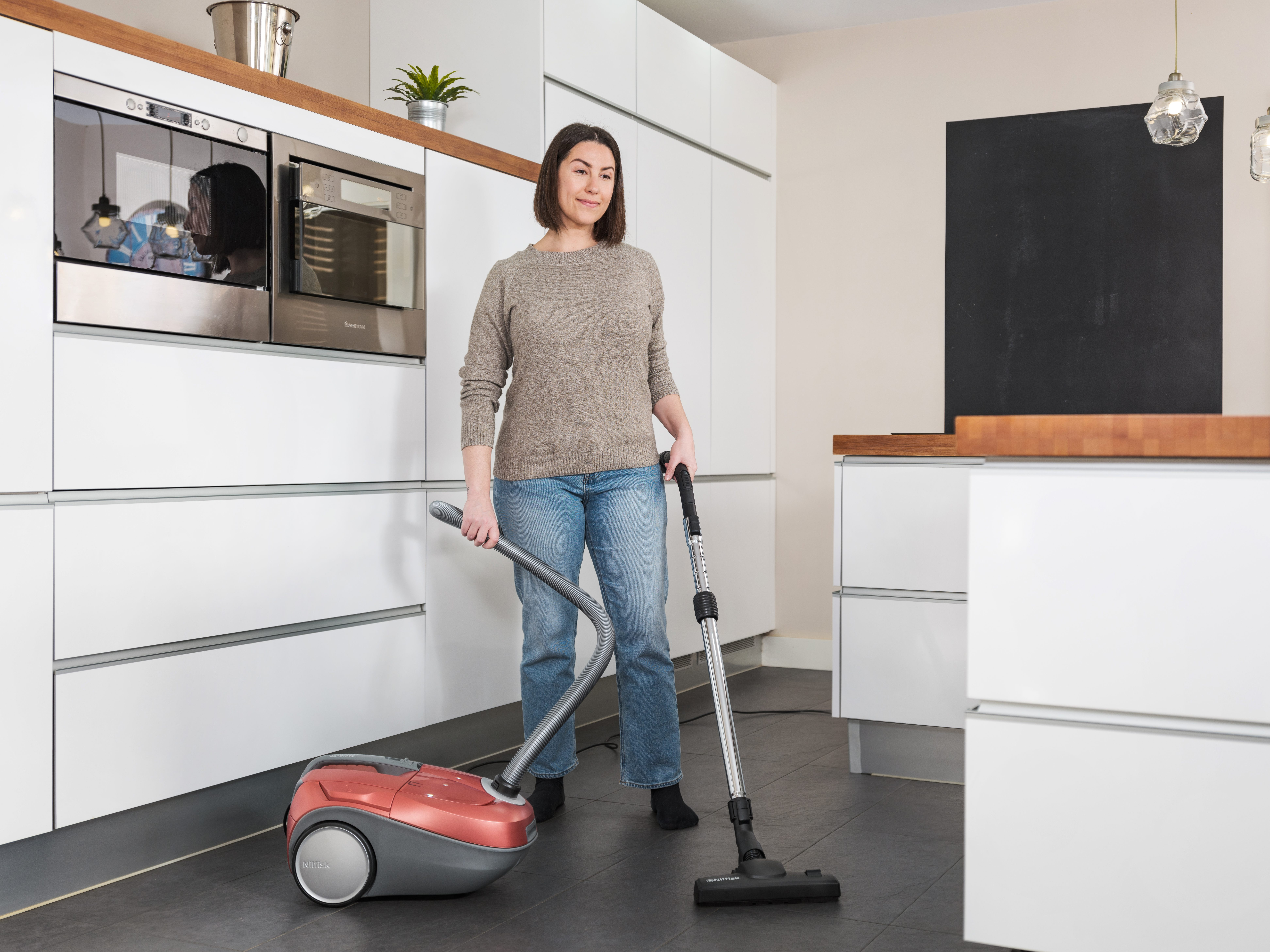 Kobieta odkurza nowoczesną kuchnię. Ma na sobie dżinsy i sweter. Widoczny jest czerwony odkurzacz.