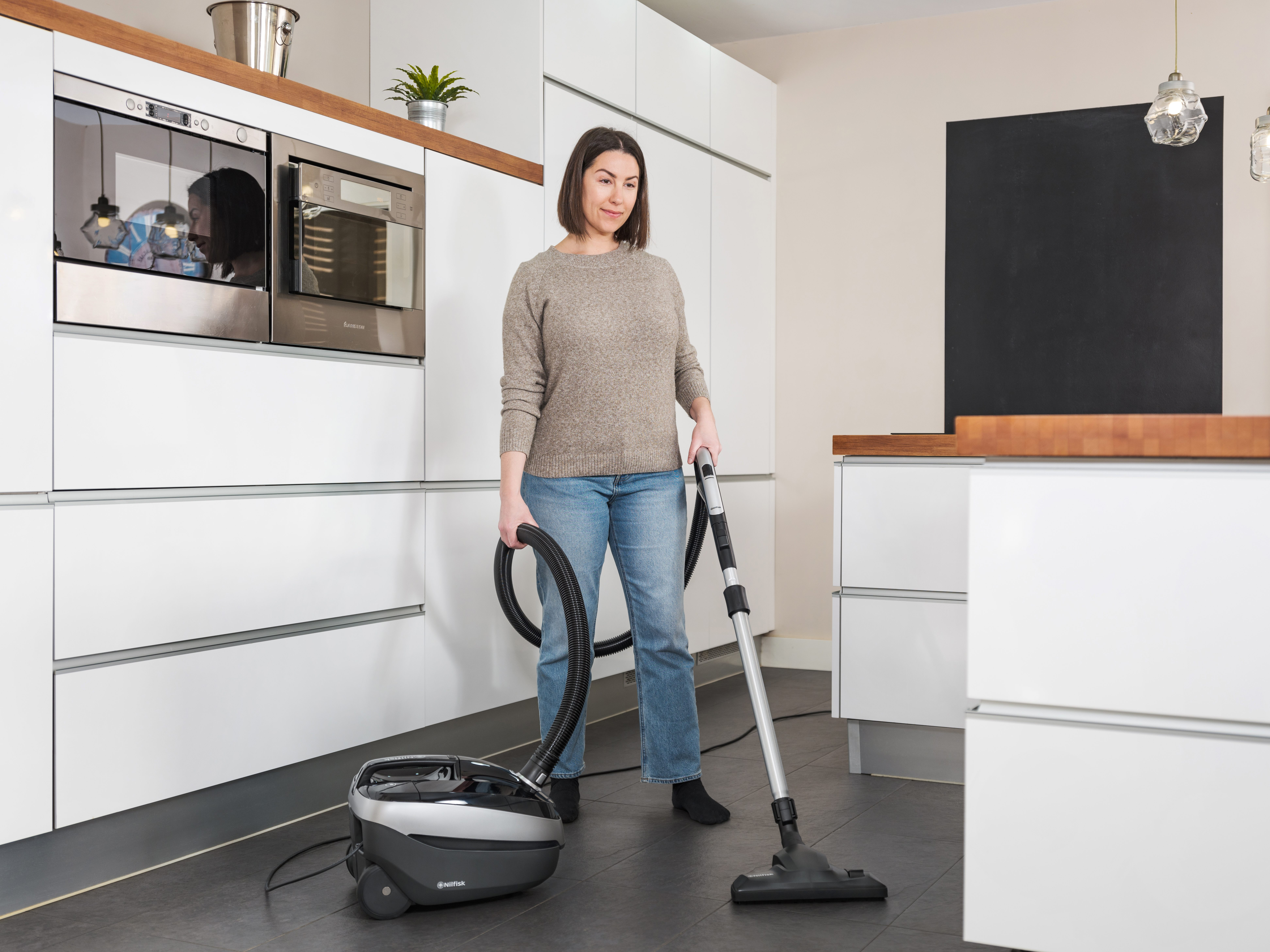 Vrouw stofzuigt een keuken. Ze draagt een spijkerbroek en een trui. Een stofzuiger staat op de grond. Witte kasten op de achtergrond.