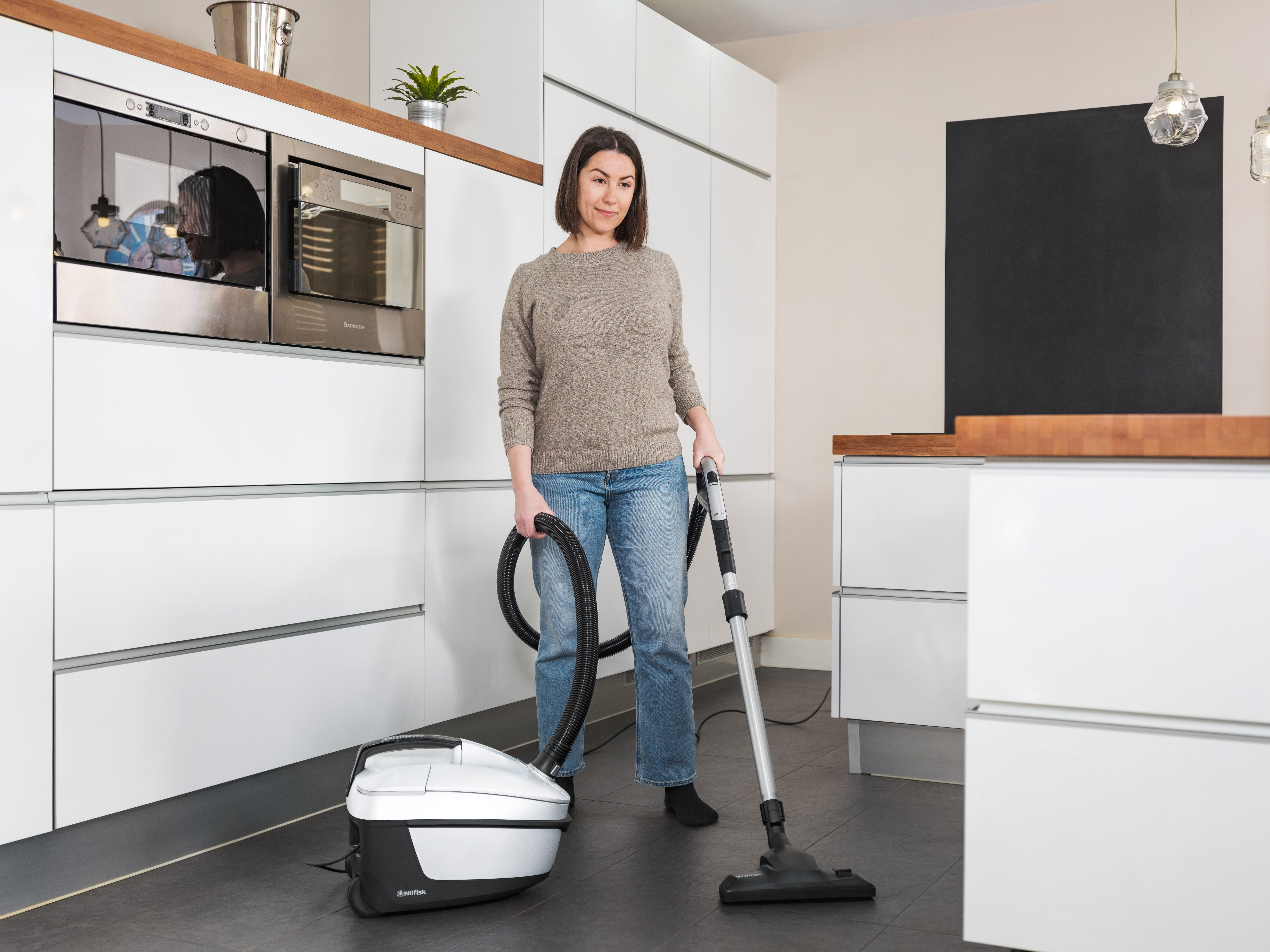 Una mujer aspira el suelo de la cocina. Sostiene la manguera de la aspiradora. Hay armarios y electrodomésticos blancos detrás de ella.