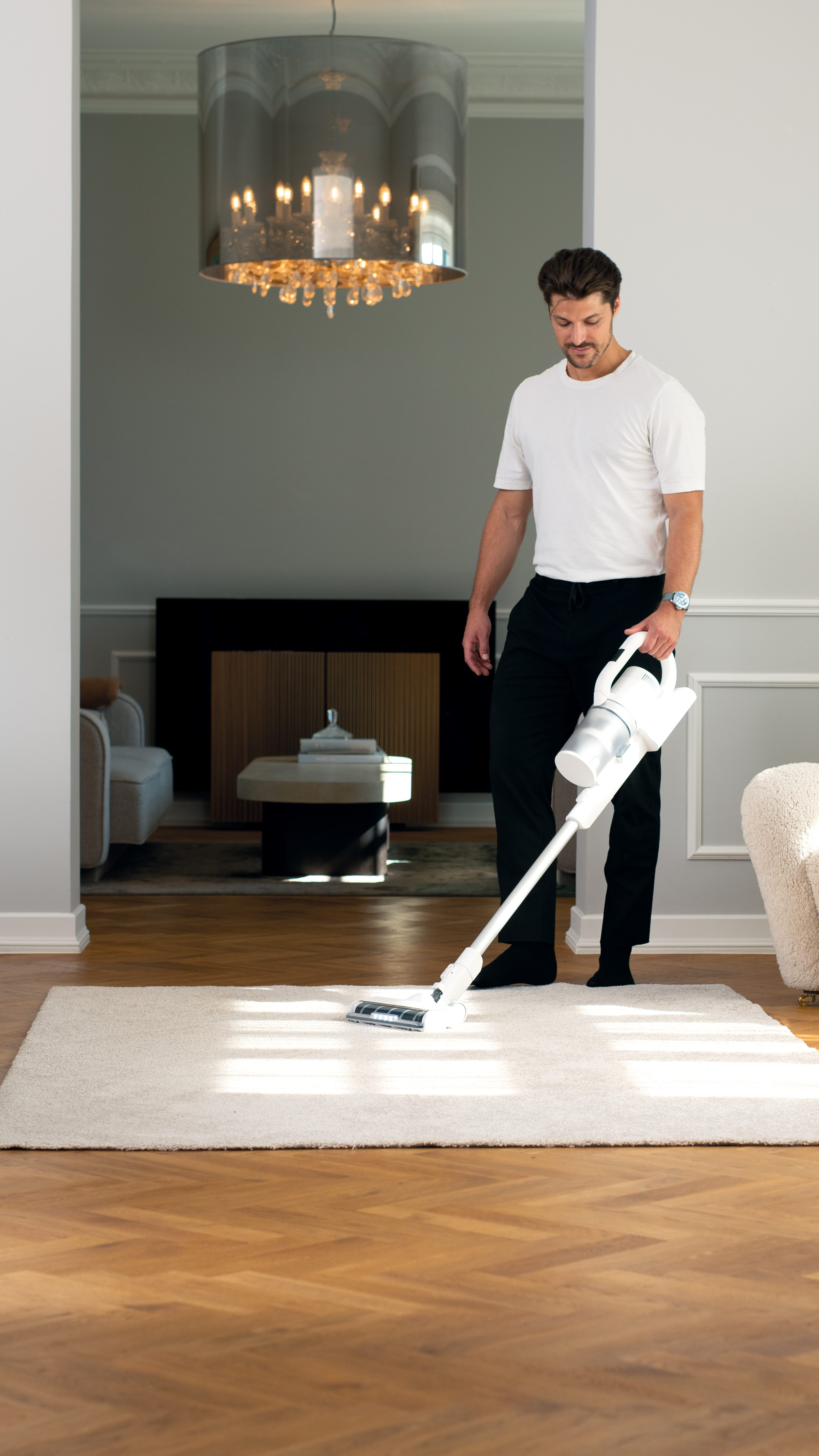 A man in a white t-shirt and black pants vacuums a light-colored rug in a modern home with a large chandelier.