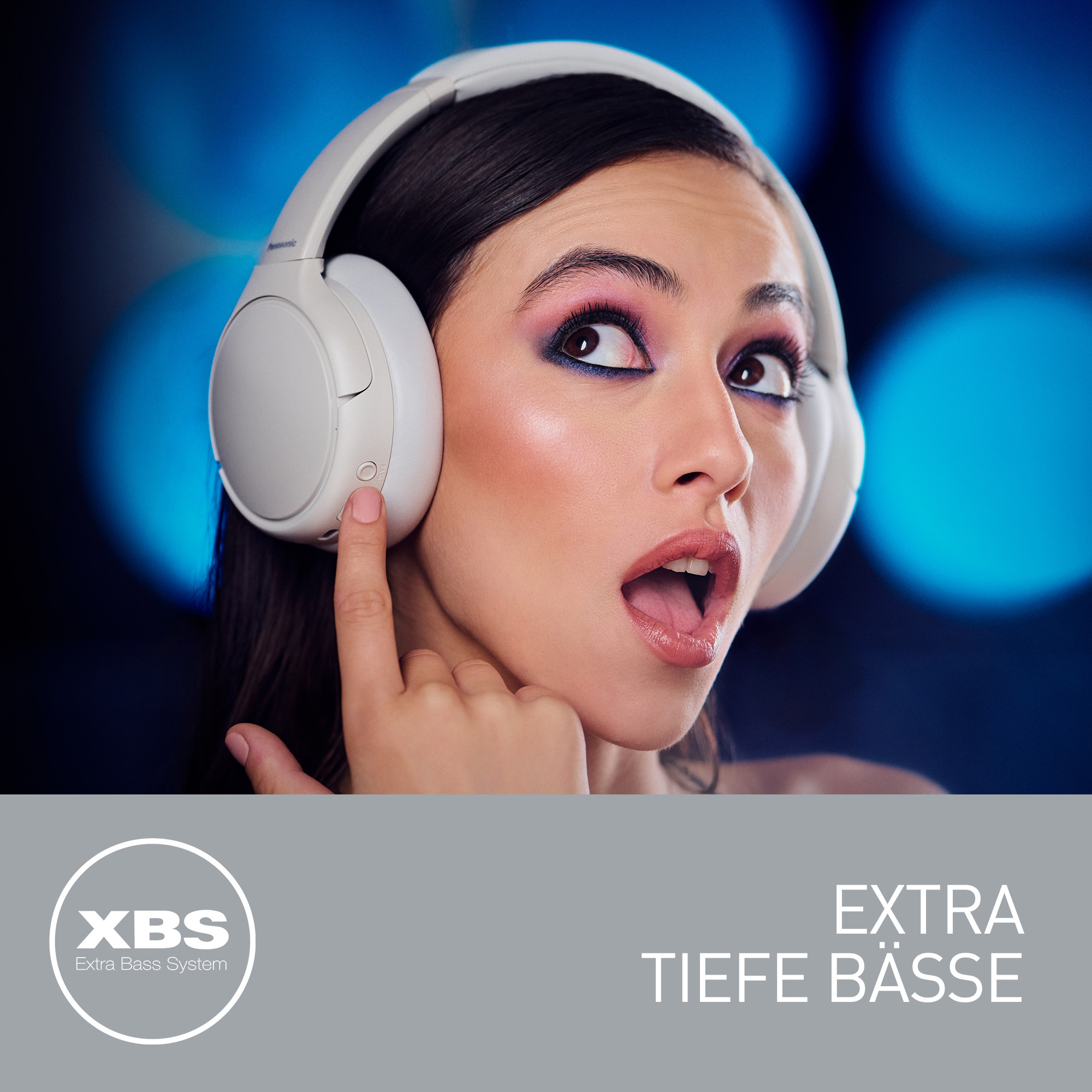 Kobieta ze słuchawkami, zaskoczona mina. Palec wskazujący na słuchawki. Tło jest niebieskie ze światłami bokeh.