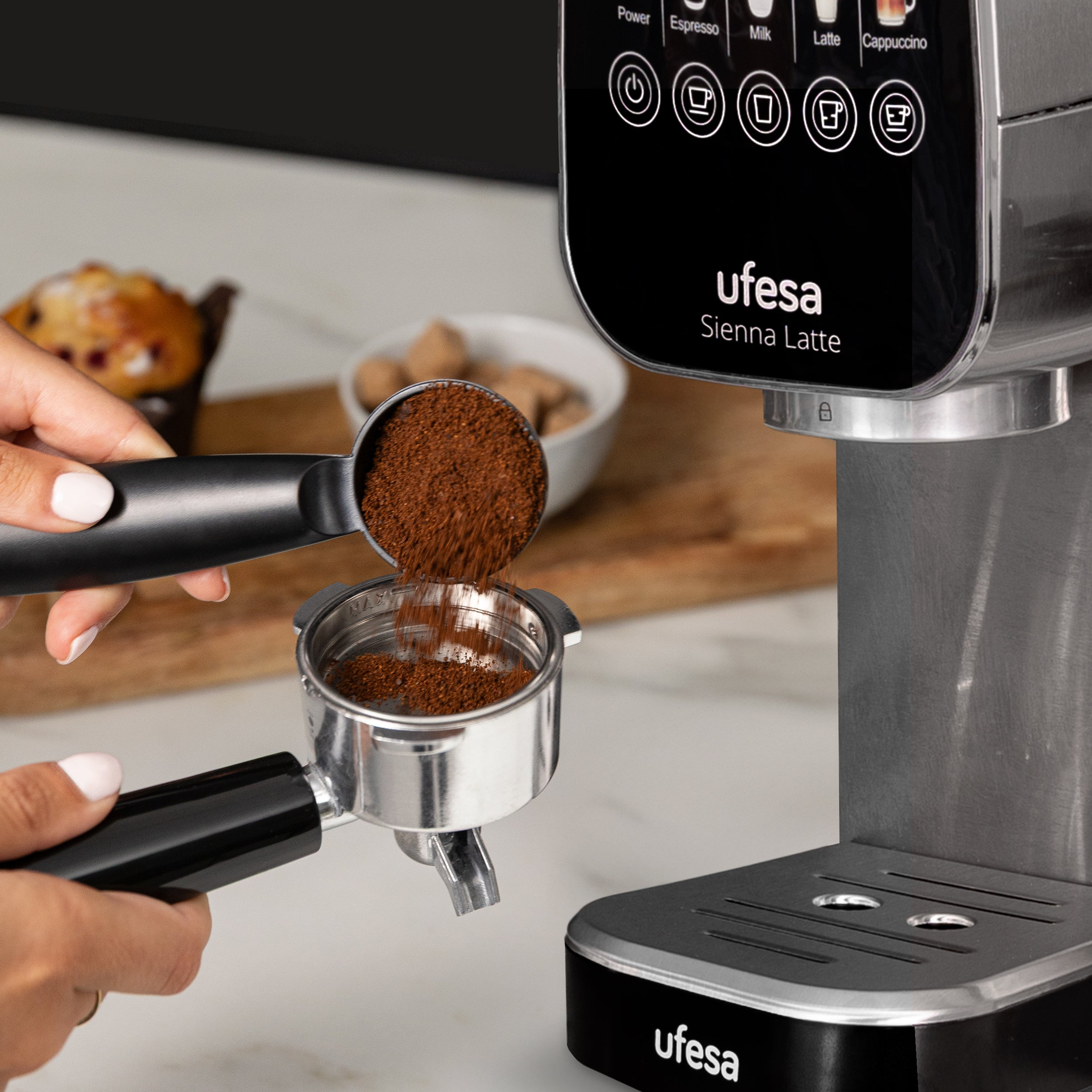 Una persona vierte café molido en un portafiltro. Una máquina de espresso está a la derecha. Un muffin en el fondo.